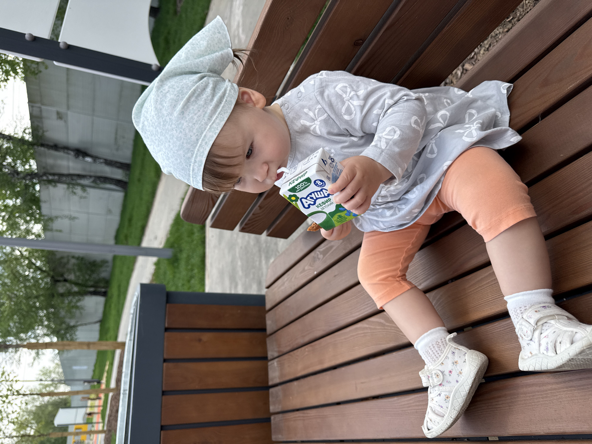 A child sits on a wooden bench outdoors. The child has light skin, brown hair, and is wearing a light blue head covering, a gray dress with white bow designs, orange shorts, white socks, and white floral sandals. The child is drinking from a small, green and white carton of "Ayusha" kefir, a dairy product. The bench is made of dark brown wooden planks. In the background, there are trees and green grass. There is a structure with white panels and dark gray supports. The location is Moscow, Russia.