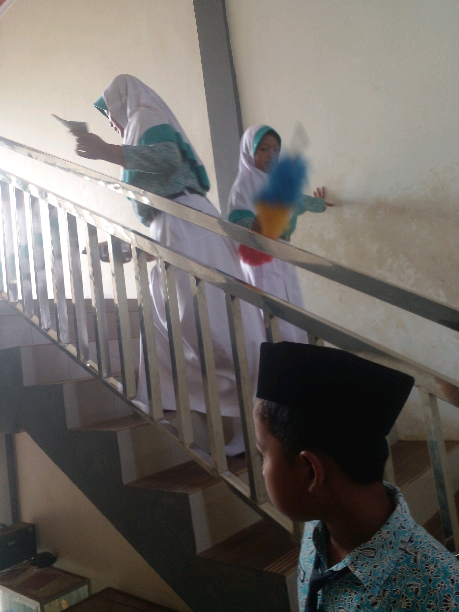 The image captures an indoor scene with a staircase. In the foreground, a young boy wearing a black cap and a blue batik shirt is partially visible, facing to the left. He appears to be looking towards the staircase. On the staircase, two girls in white and teal headscarves are present. The girl on the lower part of the staircase is holding a white piece of paper. Further up the stairs, another girl is holding a blue and yellow duster. The staircase has stainless steel railings and appears to be in a building with light-colored walls. The overall lighting suggests it is daytime, with sunlight streaming in from the left, creating lens flare. The location is identified as the city of Banyuputih, Indonesia.