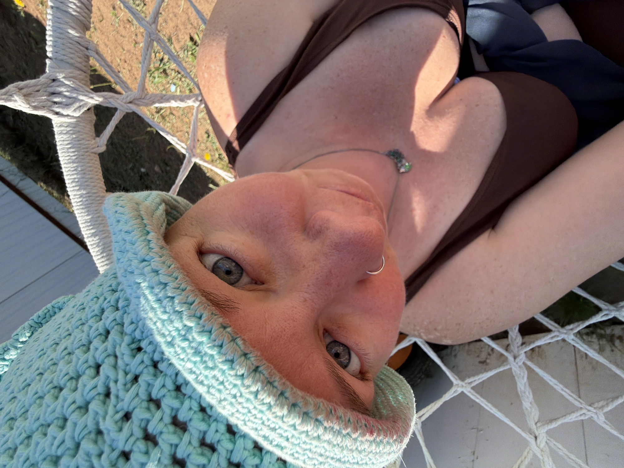 A woman with fair skin, freckles, and gray eyes is lying in a white rope hammock. She is wearing a brown top and a light blue crocheted hat. A silver nose ring is visible in her left nostril. She also wears a silver necklace with green stones. Her body is facing right and angled diagonally upwards. The hammock is outdoors, with greenery and glimpses of a white building and a fence in the background. There is no contextual information within the image that places it specifically in the city of Chaikovsky, Russia.