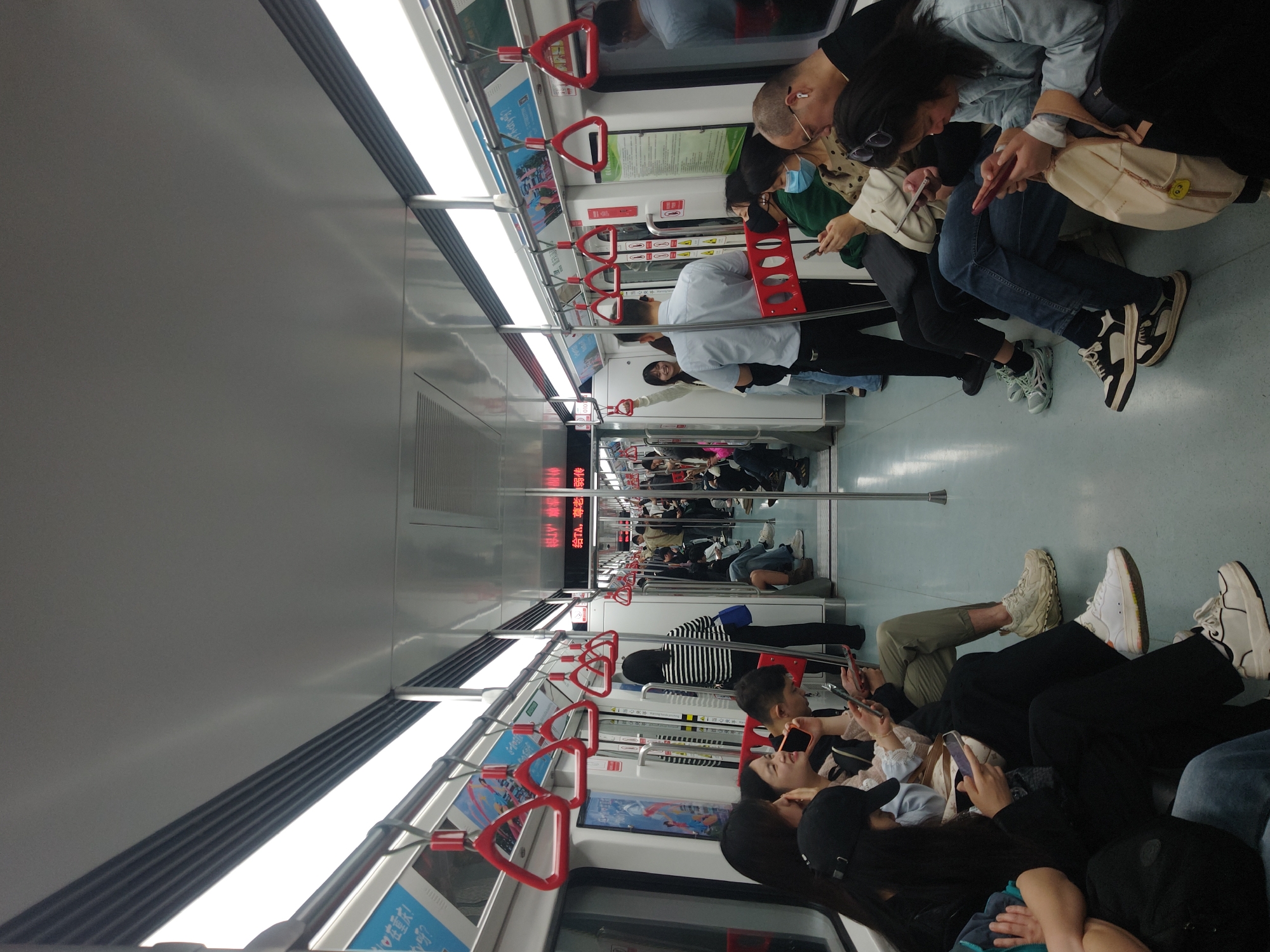 The image depicts the interior of a subway car, likely during a commute, as evidenced by the number of passengers. The car is modern, with overhead lights, metal handrails, and red triangular handholds suspended from the ceiling. Passengers are seated along the sides of the car, many of them looking at their mobile phones. A few people are standing. The overall atmosphere seems relaxed. Digital signage is visible at the end of the car. The location is not specified.