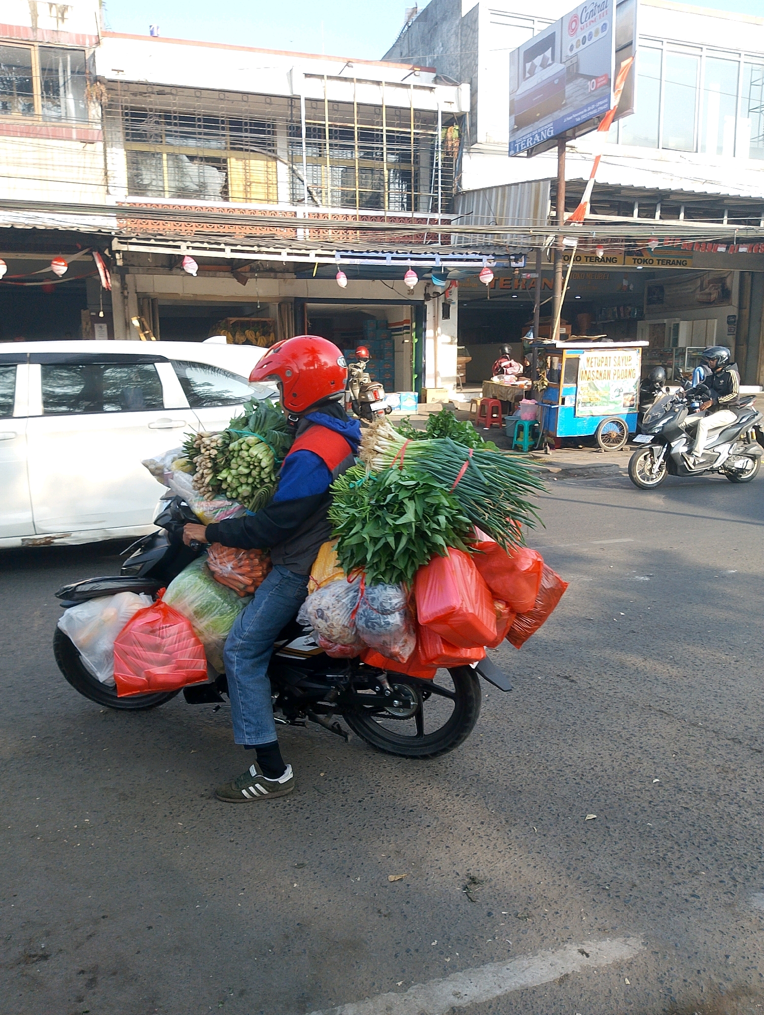 A person wearing a red helmet and a blue and red jacket is riding a black motorcycle loaded with fresh produce. The motorcycle is packed with large bundles of green vegetables, including what appears to be scallions and other leafy greens, secured in plastic bags and netting. The rider is seated on the motorcycle, facing to the right of the frame, with their legs bent and feet resting on the footpegs. They are wearing blue jeans and dark athletic shoes. The motorcycle is on a paved road, with a white car visible to the left and a food stall with a blue cart and signage in the background to the right. The scene appears to be in an urban or semi-urban environment, likely Lembang, Indonesia, given the presence of commercial buildings and a food stall. The time of day appears to be daytime, with bright sunlight casting shadows.