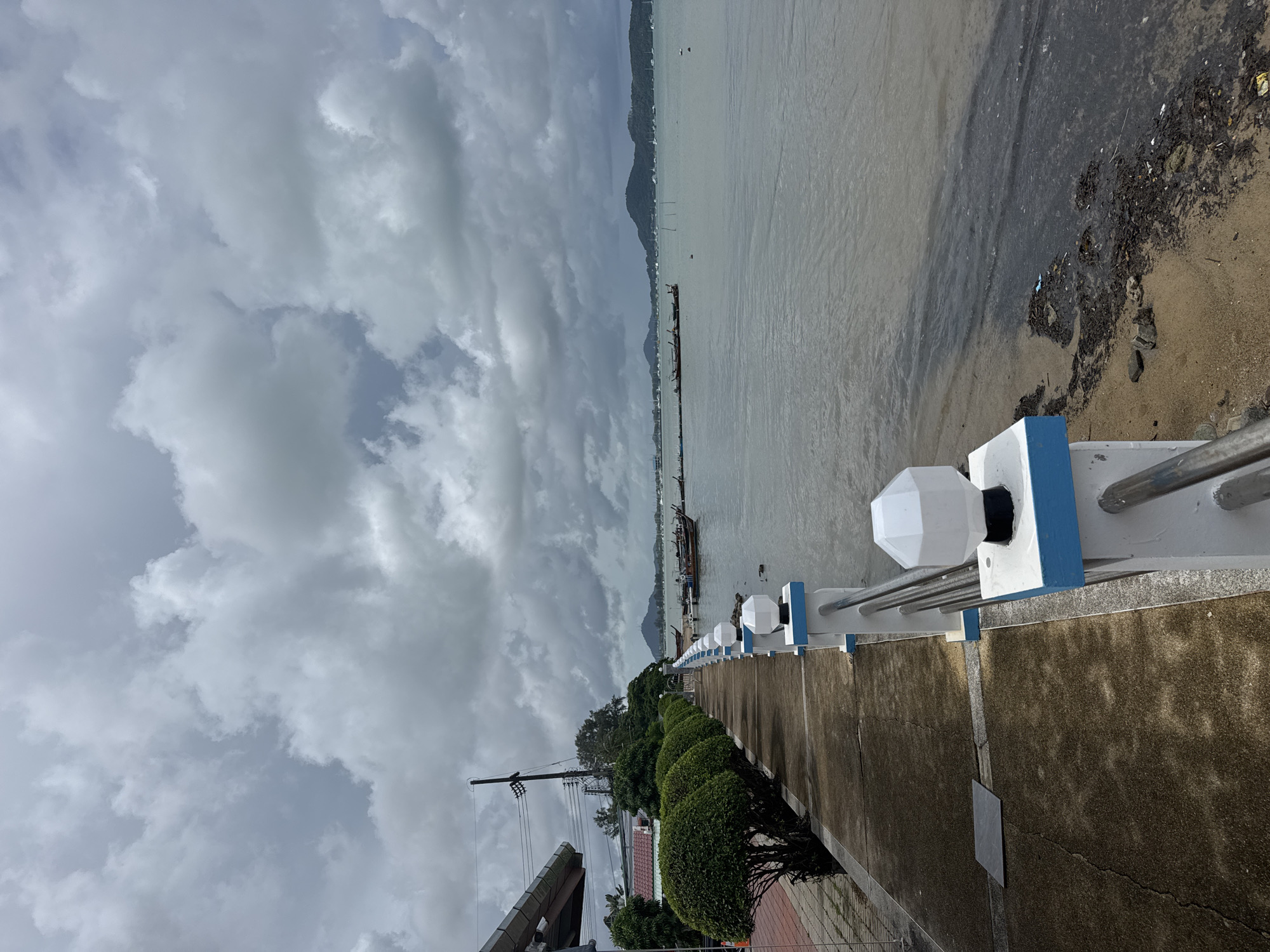 The image depicts a waterfront promenade in Rawai, Thailand, under a heavy, overcast sky. The promenade runs along the edge of a body of water, likely the sea, with a sandy beach and scattered debris visible at the water's edge. A white railing with blue accents and modern-style lamp posts lines the promenade, separating it from the water.
To the left of the promenade, there is a paved walkway and a row of meticulously trimmed, round bushes. Beyond the bushes, a building with a slanted roof and utility poles are visible. The water is a murky, gray-green color and appears relatively calm. Several traditional Thai longtail boats are moored in the water, some closer to the shore and others further out. In the distance, a mountainous island range is faintly visible on the horizon, with a scattering of smaller boats or yachts. The overall atmosphere is somber due to the cloudy weather.