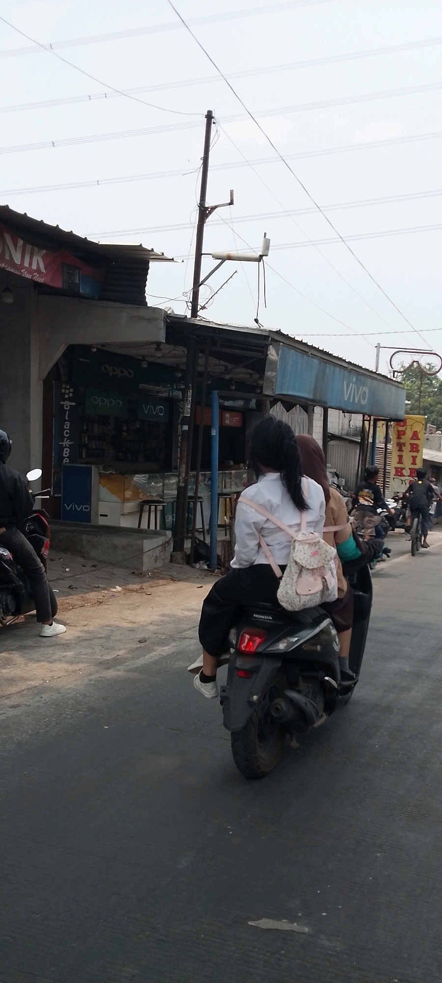 Two young women are riding on a motorcycle on a street in the city of Bekasi, Indonesia. The woman in the front is wearing a white collared shirt, black pants, and white sneakers. She has her dark hair tied back in a ponytail and is carrying a small, floral-patterned backpack. The woman behind her is wearing a brown hijab and a brown shirt. They are on a dark-colored motorcycle.
The background shows a street with several small shops or stalls. One stall has signs for "OPPO" and "VIVO," suggesting it sells mobile phones. There are utility poles and power lines overhead, and the sky is overcast. Other people and motorcycles are visible in the distance, indicating a busy street. The overall scene depicts a typical street view in an urban area of Indonesia.