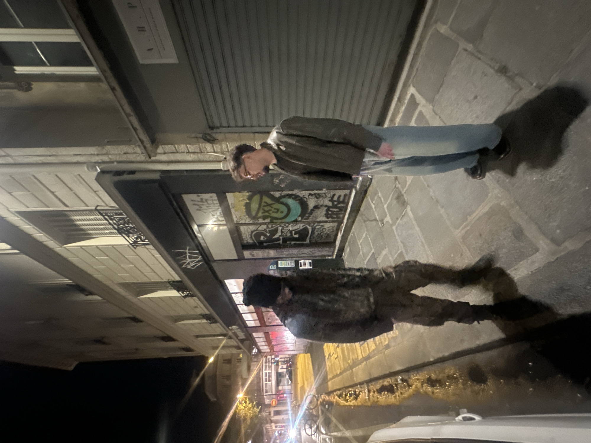 The image captures a street scene in Paris at night. The camera is tilted, giving a distorted perspective. Two figures are present: one standing on the sidewalk in the upper part of the image, wearing jeans and a jacket; the other is positioned lower, possibly standing on the street, wearing a darker outfit and a cap. In the background, there is graffiti-covered storefront and a view of buildings and streetlights receding into the distance. A car is partially visible in the lower part of the image. The sidewalk and road surface suggest a typically urban environment.