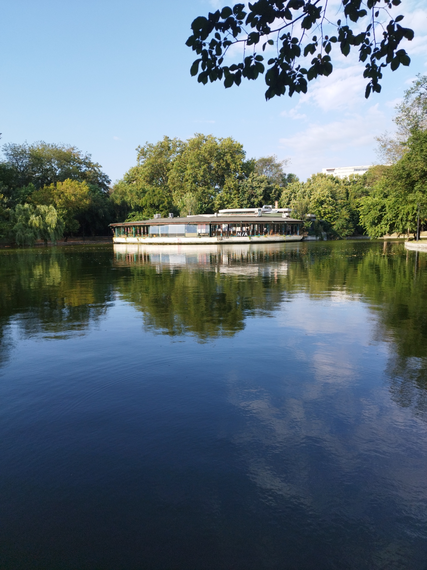 A circular, glass-walled restaurant on a lake in Bucharest, Romania. The restaurant, named "N/A," is surrounded by trees and reflects in the calm water. The sky is clear blue with a few scattered clouds.