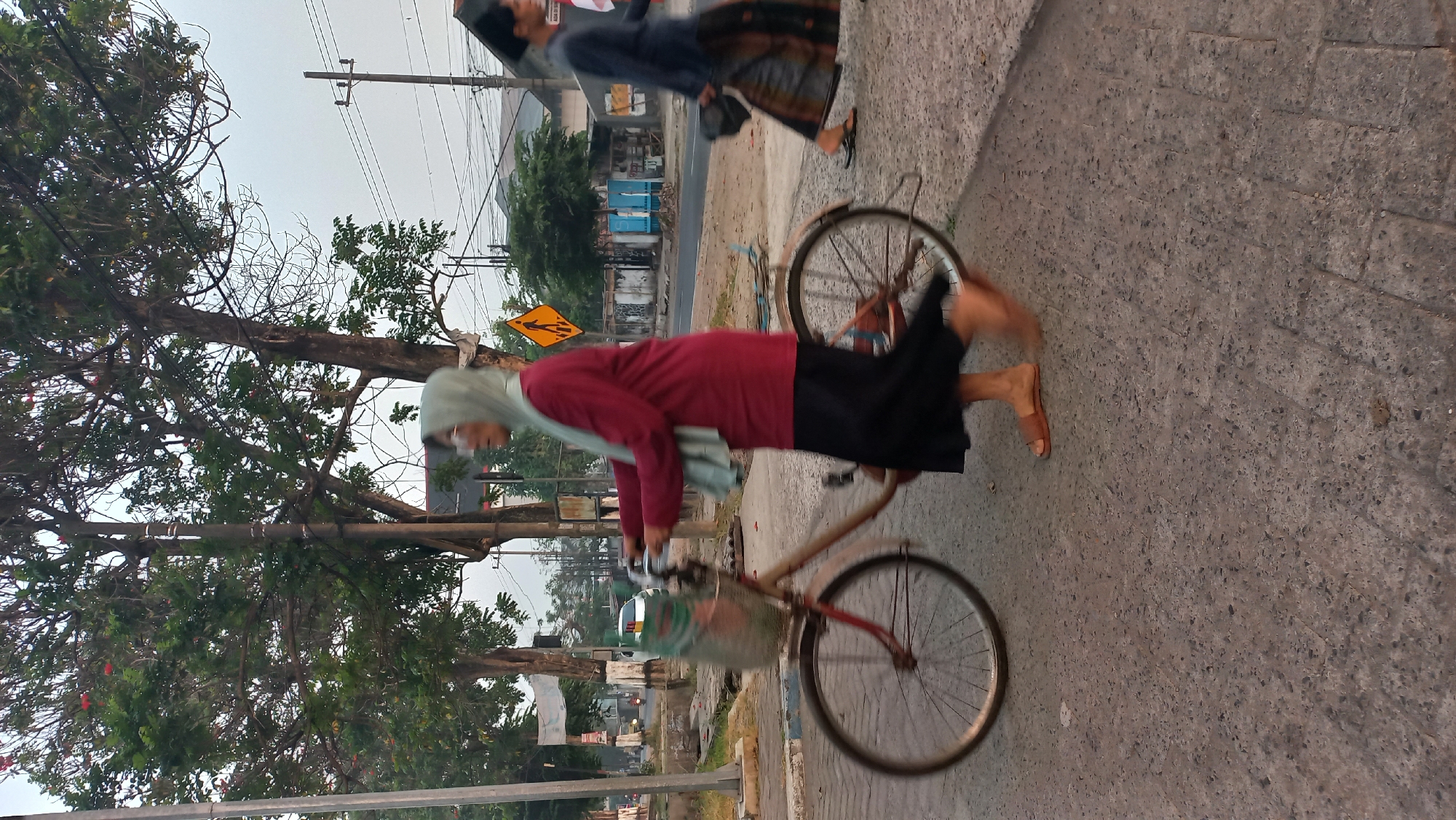 The image captures a street scene in Pare, Indonesia, likely in the morning or late afternoon given the soft lighting. A woman wearing a light blue hijab and a maroon long-sleeved shirt with a black skirt is pushing a bicycle. The bicycle has a basket on the front, which appears to contain some items covered by a green mesh bag. The woman is wearing brown sandals. Her motion is blurred, suggesting she is walking or moving the bicycle forward.
To the right of the woman, a person wearing a dark blue shirt and a patterned sarong is walking away from the camera, also with blurred motion. This person is carrying a dark object, possibly a hat, in their right hand.
The environment includes a paved road with some visible traffic lights and power lines overhead. Trees with green foliage are present on the left side of the frame, and further down the street, buildings and more trees are visible. A yellow diamond-shaped pedestrian crossing sign is mounted on a pole in the mid-ground. The overall impression is a typical street in a town or city in Indonesia, with everyday activities taking place.