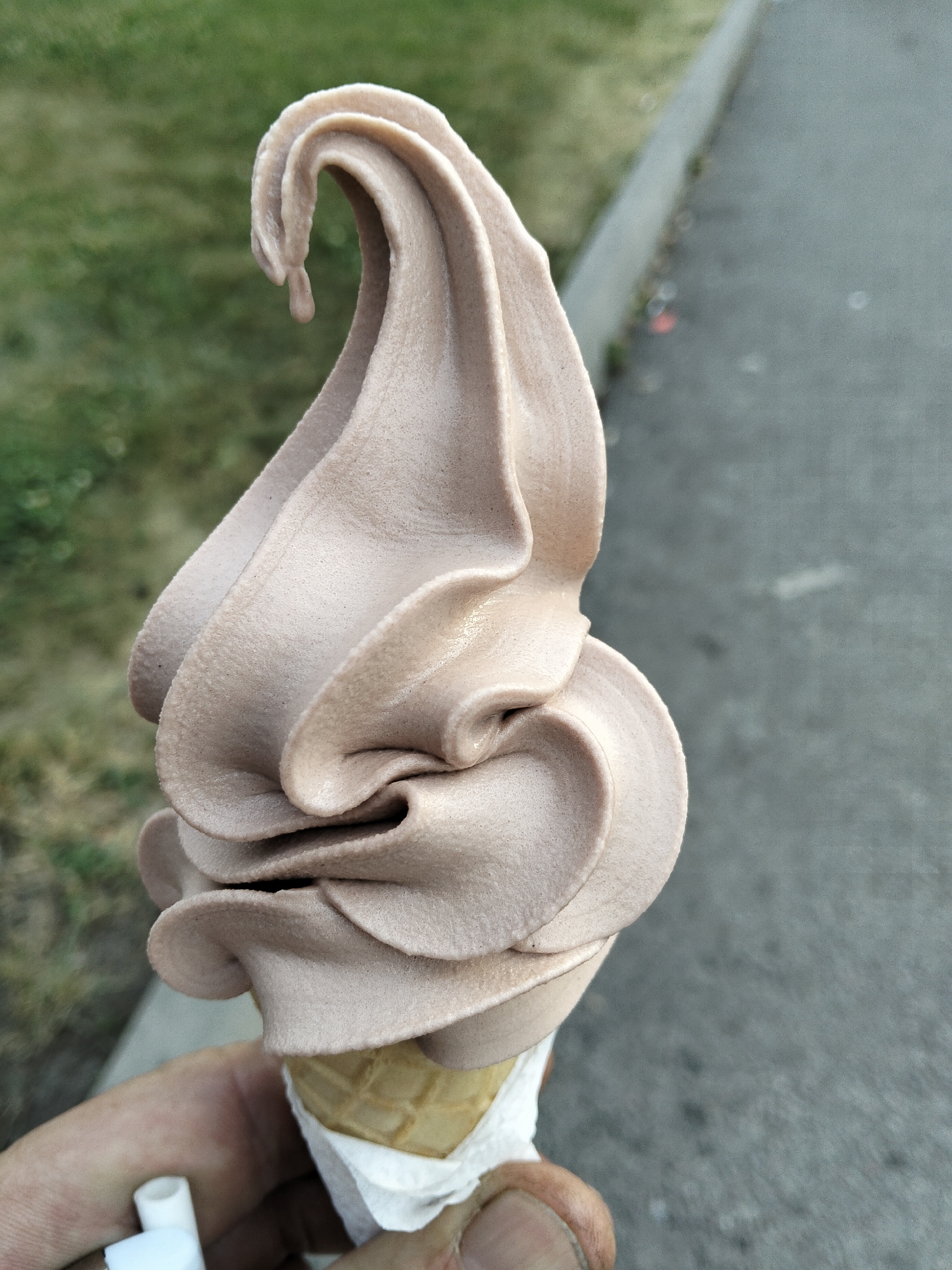 An extreme close-up shot shows a person holding a chocolate soft-serve ice cream cone. The ice cream is a light brown color and is swirled in a tall, elegant shape, with a small drip hanging from the top curl. The cone is light brown and waffle-patterned, partially wrapped in a white paper holder. The person's hand, with visible skin texture and dirt under the fingernails, is holding the cone at the base. In the background, a blurred green grassy area is visible on the left, transitioning to a grey paved surface on the right, which appears to be a sidewalk or path. The lighting suggests an outdoor, possibly daytime setting. There is no direct evidence to indicate the specific location within the city of Almaty, Kazakhstan.