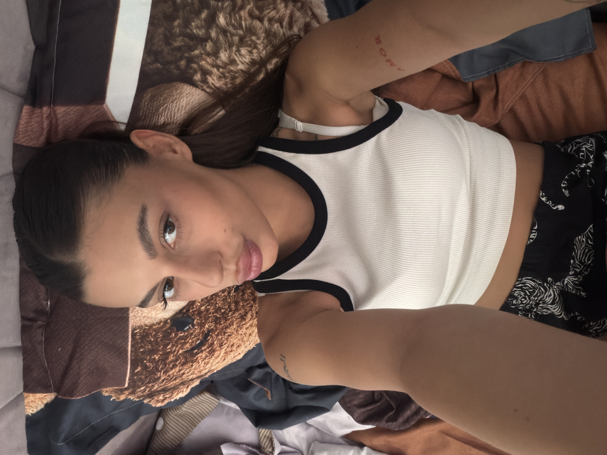 The image features a young woman lying on a bed. She is wearing a white ribbed tank top with black trim and patterned black shorts. Her hair is dark and pulled back. Tattoos are visible on her arm. The bed has a pillowcase with a teddy bear design and is covered with various fabrics, suggesting a relaxed or casual setting. The lighting appears to be natural, possibly from a window, and there are no clear indicators of the time of day or weather conditions. The woman's expression is calm, and she is looking towards the camera. There is visible text on her arm tattoo which reads "AMOR".