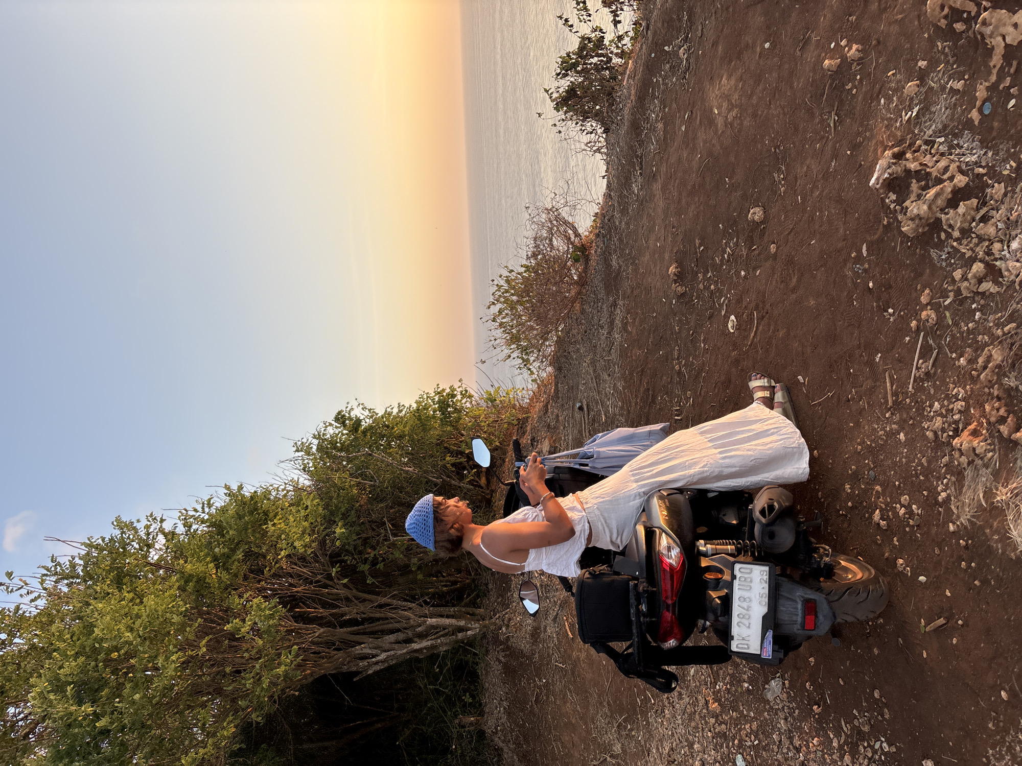 A woman wearing a white top, a white long skirt, and a blue knitted hat sits on a black scooter parked on a dirt cliff overlooking the ocean at sunset. The scooter is equipped with a black storage box on the back and a license plate reading "DK 2848 UBQ". The woman holds a phone in her hands and appears to be looking at it. A light blue bag hangs from the back of the scooter. The background consists of trees and bushes on the left, and the vast ocean meeting the pale orange and yellow sky on the right. The ground is covered in dirt, rocks, and dry vegetation. The scene is set in Kuta Selatan, Indonesia, characterized by its coastal landscape and sunset views.