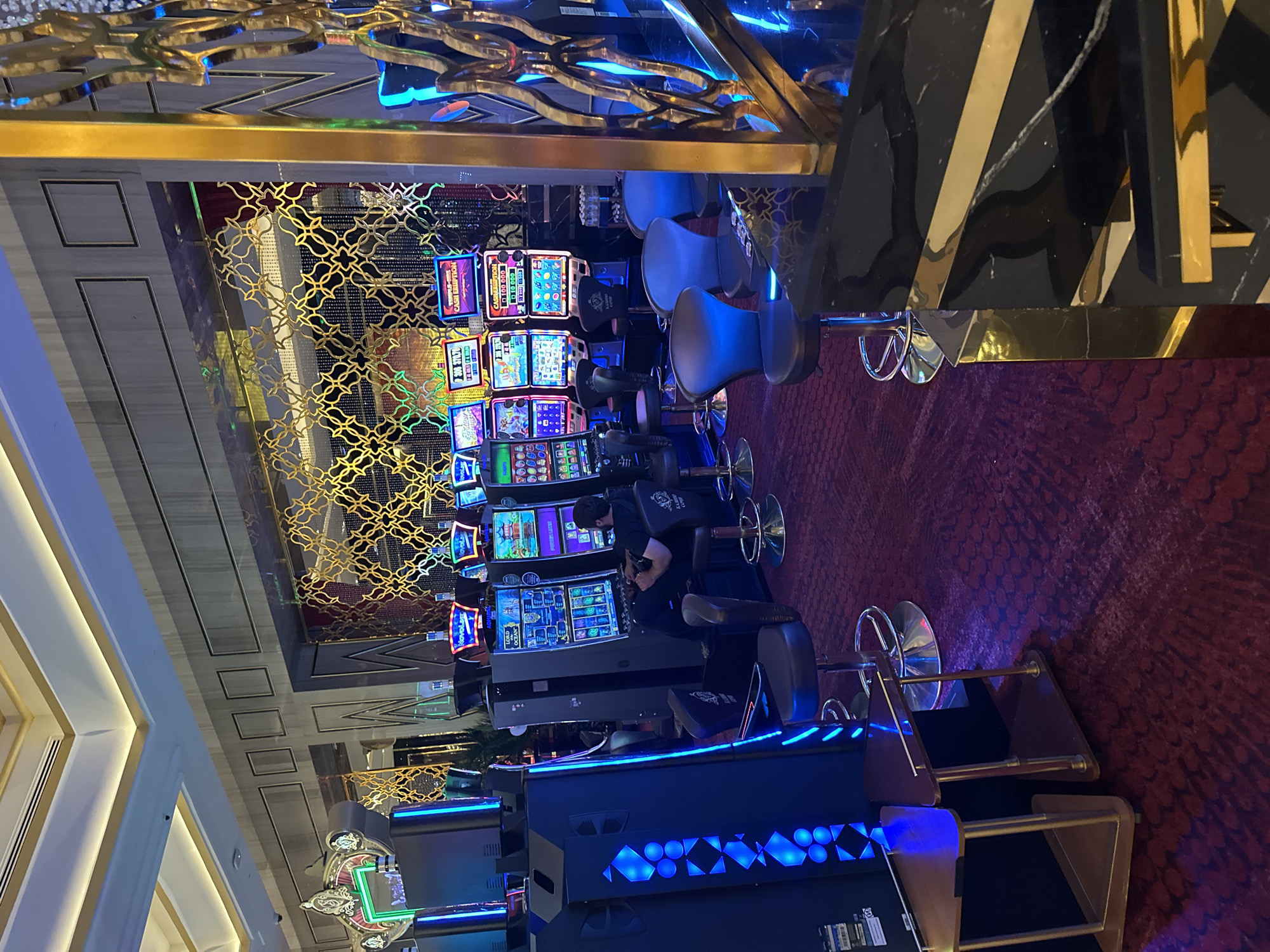 The image shows an interior view of a casino in Estosadok, Russia. The scene is dominated by rows of slot machines, many of which are lit up with vibrant colors. In the foreground, to the left, is a large, black slot machine with blue illuminated geometric patterns on its side. Next to it are two small triangular tables with gold legs. Further back, a man is seated at a slot machine, facing away from the camera. He is wearing a black t-shirt and dark pants. Several other slot machines are visible in the background, with their screens displaying various game graphics. Blue and dark chairs with silver bases are positioned in front of the machines. The environment is luxuriously decorated with ornate gold patterned screens and textured wall panels. The flooring is a deep red carpet with a patterned design. The ceiling is high and features recessed lighting and gold accents.