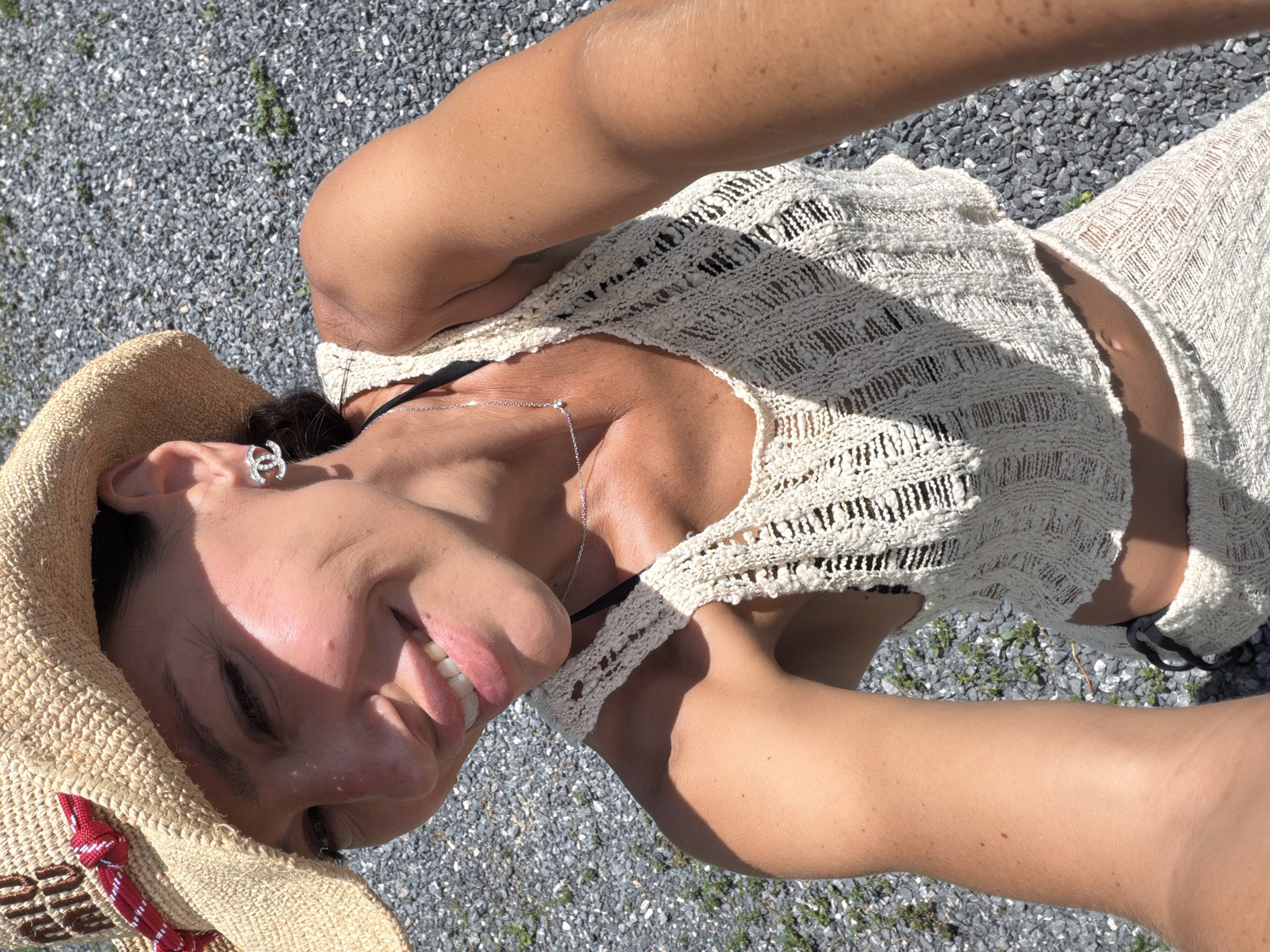In Baan Bo Phut, Thailand, a woman wearing a straw hat and a white crochet top takes a selfie outdoors on a sunny day. The background consists of grey gravel and sparse green vegetation. The woman has on a silver necklace and Chanel earrings.