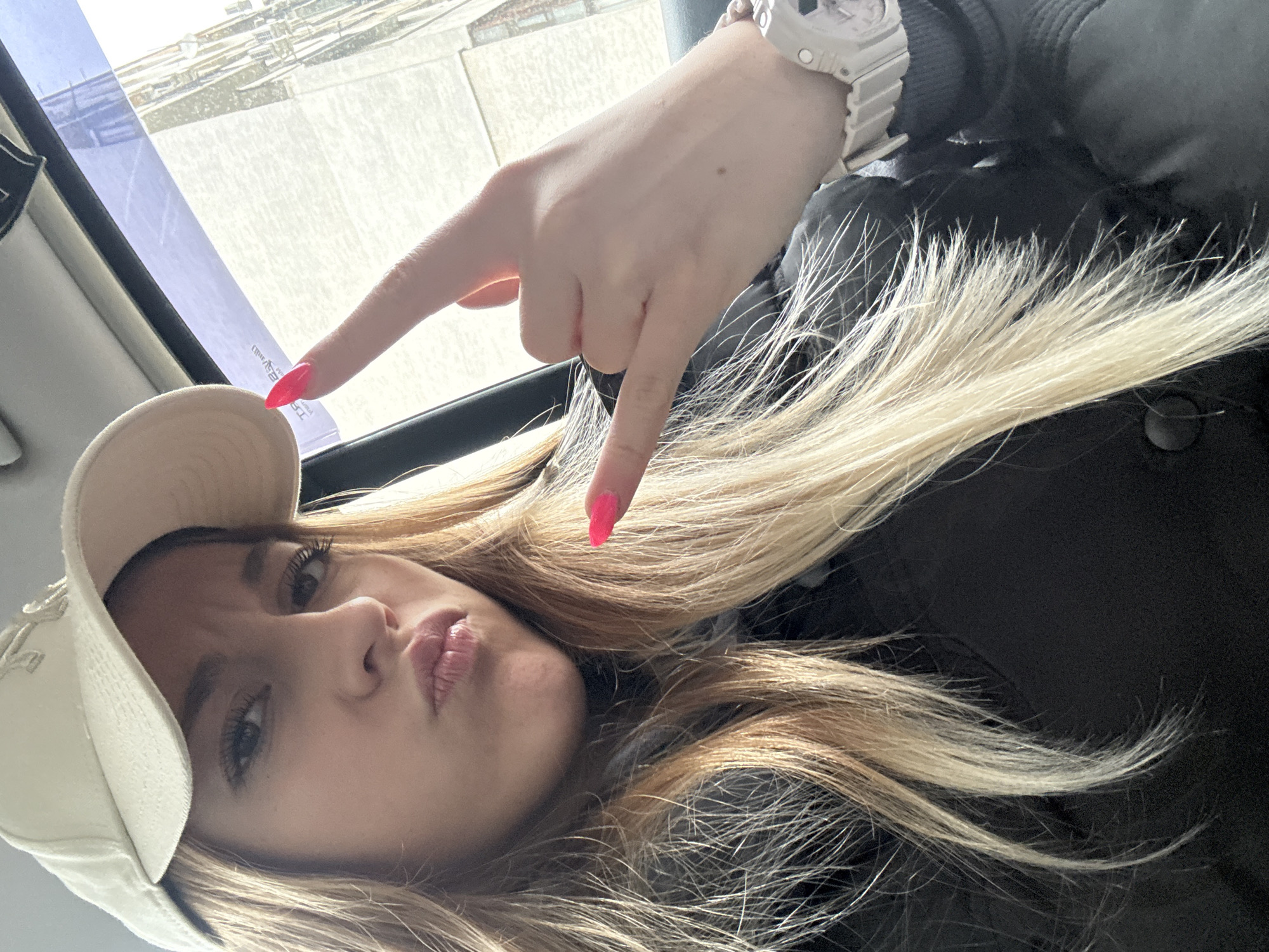 A young woman with blonde hair and wearing a white cap and watch is in the passenger seat of a car. She is making the "rock on" hand gesture with bright pink nail polish. The background shows a blurred urban setting, potentially in a city like Sofia, Bulgaria, given the architectural style visible through the window.