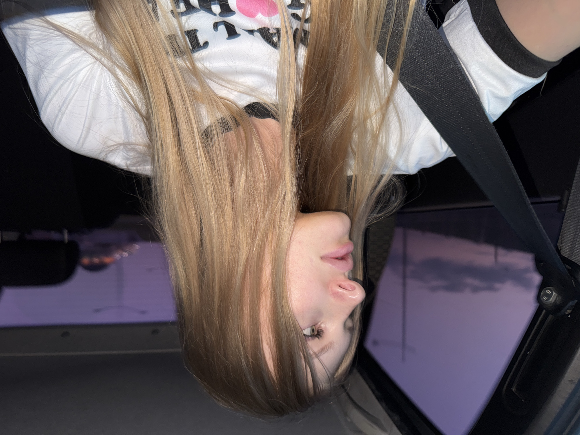 A young woman with long blonde hair is sitting in the passenger seat of a car, looking out the window at a purple sky. She is wearing a white t-shirt with black trim. A black seatbelt is across her body. The car appears to be in motion, indicated by the blurry background of city lights and streetlights in the distance. The sky suggests it is either sunrise or sunset.