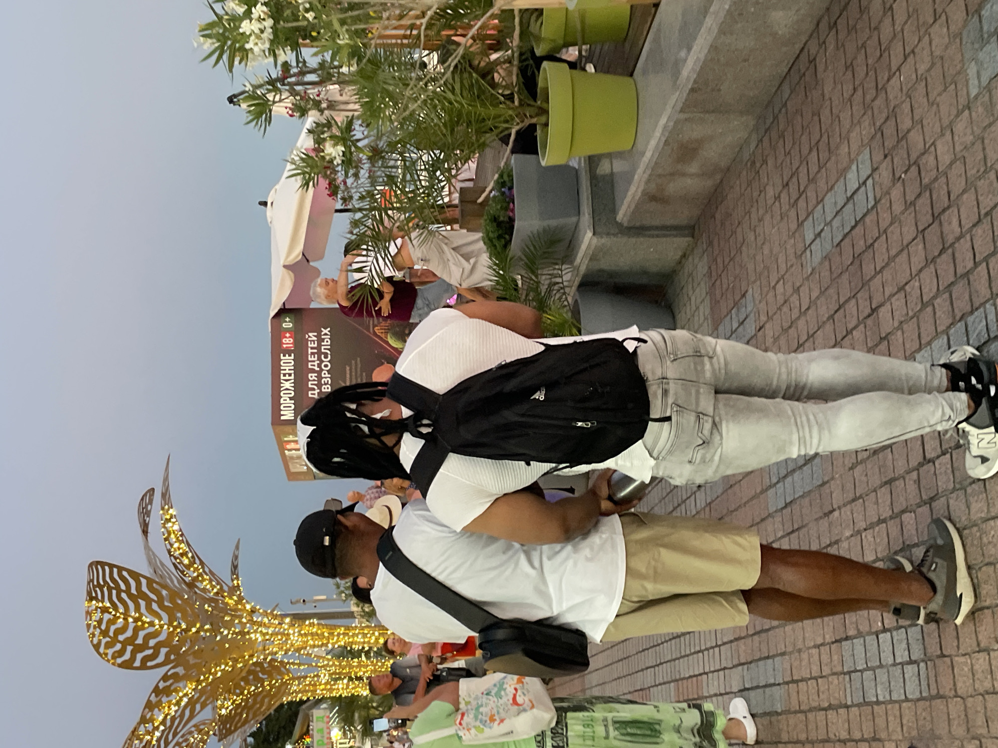 The image depicts a street scene in Sochi, Russia, possibly during the evening or late afternoon, given the twilight sky and decorative lighting. In the foreground, two individuals with backpacks are facing away from the camera, walking on a paved walkway. The person on the left wears a white t-shirt, khaki shorts, and grey sneakers. The person on the right wears a white t-shirt, light-colored jeans, and sneakers. Behind them, a large, stylized, illuminated golden tree structure is visible, suggesting a festive or decorative element in the urban environment. Further in the background, there is a stall with signage that appears to advertise "ICE CREAM 18+" in Russian, and mentions "FOR CHILDREN ADULTS." Several other people are scattered throughout the scene, some interacting and others walking. Lush green plants in pots, including palm trees and flowering bushes, are present, adding to the outdoor ambiance. A white umbrella is also visible, possibly part of a cafe or seating area. The overall impression is of a public space, likely a promenade or pedestrian area, with commercial activity and people enjoying the outdoors.