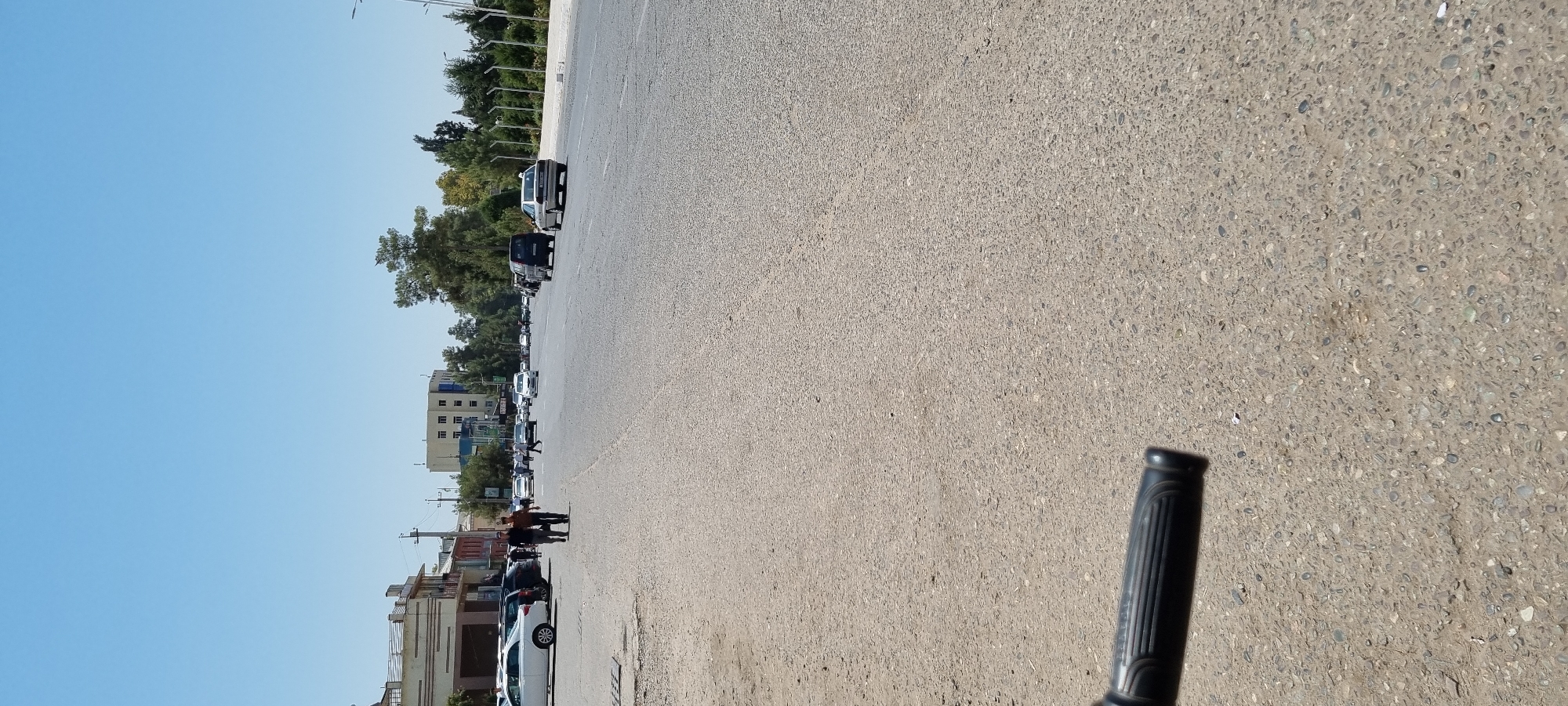 The image depicts a street scene in Ismoili Somoni, Tajikistan, on a clear, sunny day. The foreground shows the textured asphalt of a road and a portion of a black handlebar, suggesting the perspective is from a bicycle or similar vehicle. In the middle ground, several cars are present, including a white sedan, a dark-colored van, and a silver sedan. Two men are walking on the sidewalk on the left side of the road. Further down the street, more vehicles and pedestrians are visible, and buildings line the left side. The background features a clear blue sky, trees, and larger buildings, including a multi-story structure with visible windows. The scene appears to be an active urban environment with moderate traffic.