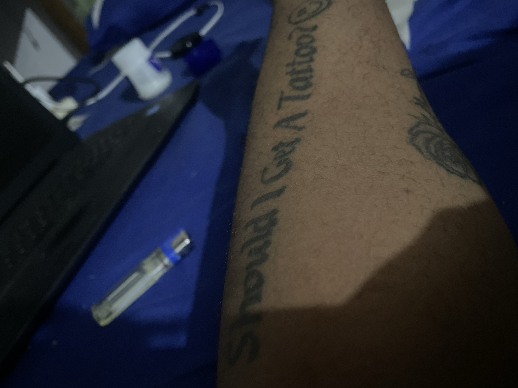 A close-up shot of a person's arm with a tattoo that reads, "Should I Get A Tattoo? 🤨". The tattoo is in black ink, and the skin tone is brown. The arm rests on a blue surface. To the left, there is a black laptop, a clear lighter with a blue cap, and what appears to be a charging cable and adapter. The background is a mix of blue and white, likely part of bedding or other items. The city of Jos, Nigeria, is not discernable from the visual information provided.