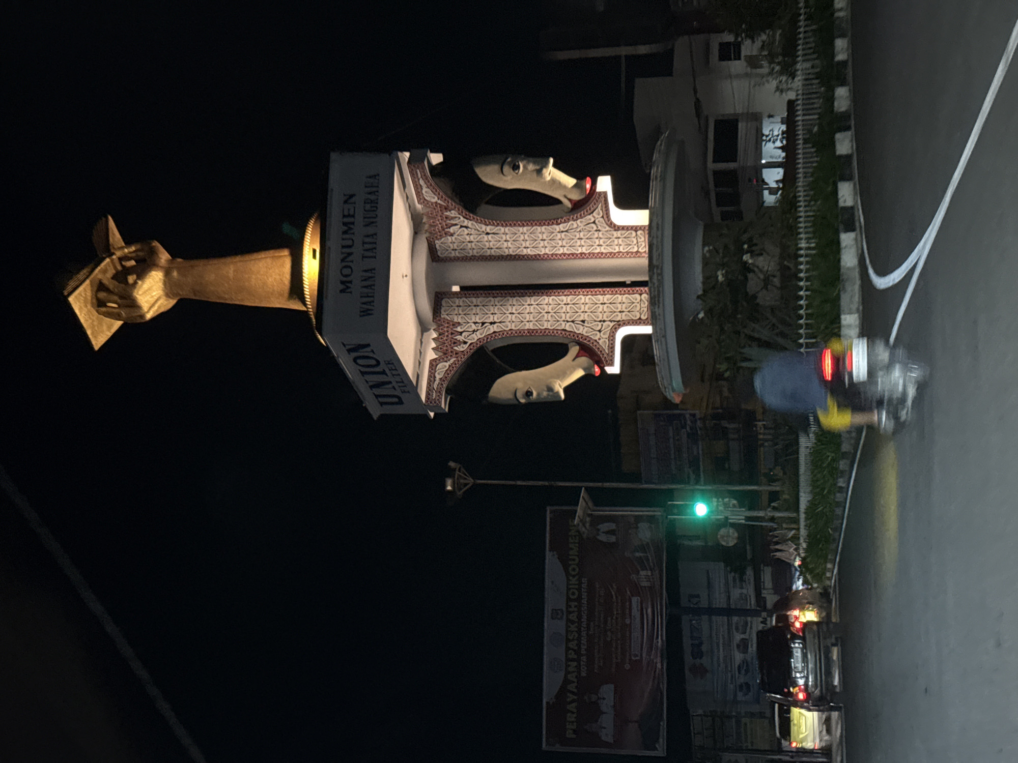 This is a nighttime, outdoor shot in the city of Pematang Siantar, Indonesia. The image features a large, ornate monument with decorative carvings and two prominent faces. To the left of the monument, a golden-colored hand holds an object that resembles a book or tablet. On the monument itself, text reads "MONUMEN WAHANA TATA NUGRAHA" and "UNION FILTER".
In the background, a street is visible with a traffic light showing green. Several cars are present on the road, some with their headlights on. A person is seen riding a scooter or motorcycle on the right side of the image, with yellow shorts and a blue shirt. The setting appears to be urban, with buildings and streetlights visible. A sign with text including "PERAYAN PASKAH OIKUMENE" and "KOTA PEMATANGSIANTAR" is also visible. The overall mood is dark due to the nighttime setting, with artificial lighting illuminating the monument and surrounding areas.