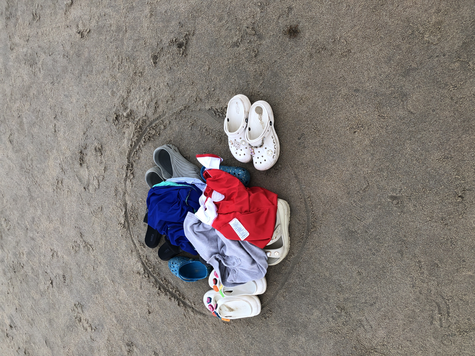 A pile of clothing and footwear sits on a sandy beach, likely in Đà Nẵng, Vietnam, within a circular imprint in the sand. The clothing consists of a blue shirt or jacket, a gray long-sleeved shirt, and a red shirt. Various types of footwear are present, including several pairs of white slip-on sandals adorned with floral designs and charms, a pair of blue glittery clogs, two pairs of black flip-flops, and at least two pairs of light gray slip-on shoes or clogs. The items are loosely arranged, suggesting they have been recently removed or are about to be put on. The background is a textured sandy beach with faint impressions and debris scattered across the surface.