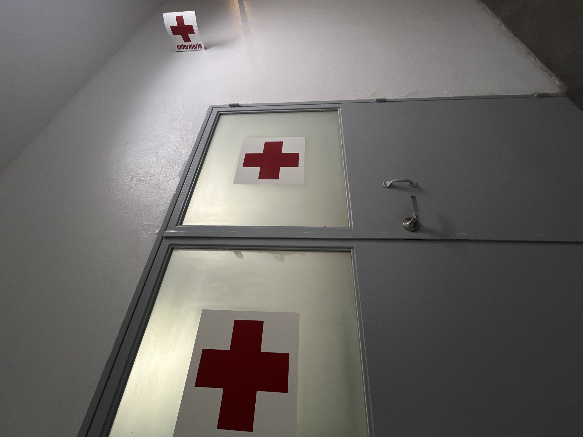 The image shows a door with a red cross symbol on each of its two glass panels. The door is likely for a medical facility. Above the door, on the wall, is a sign with the word "enfermería" in Spanish and a red cross symbol. The walls and door are painted a light gray color. The location is unknown. There are no people visible in the image.
