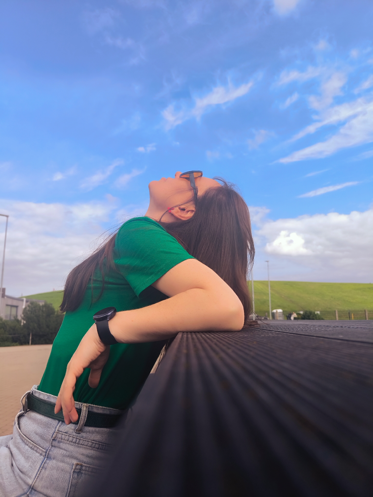 A young woman with long dark hair leans back against a dark-colored railing, her head tilted back, looking up at the sky. She is wearing a green t-shirt, light-wash jeans, a dark belt, and sunglasses. A black smartwatch is visible on her left wrist. The background features a bright blue sky with scattered white clouds, a grassy hill, some buildings, and trees. The image appears to be taken outdoors in an urban or suburban setting, possibly in Riga, Latvia, given the architecture and landscaping.