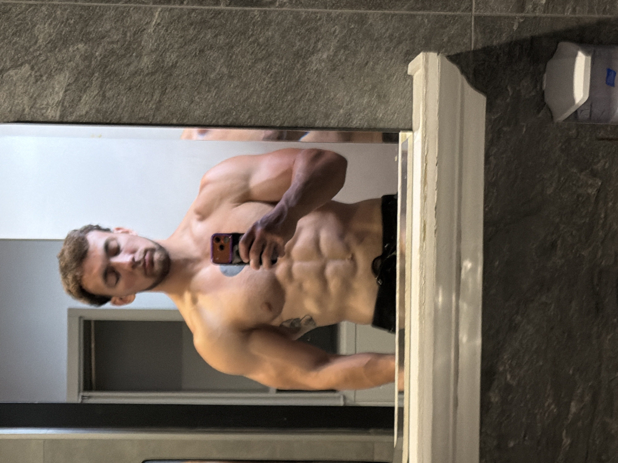 The image shows a man taking a mirror selfie in a bathroom. He is shirtless, revealing a muscular physique with well-defined abs and arms. He is holding a phone in his right hand, which is pointed at the mirror. The background of the bathroom includes a gray tiled wall, a doorway, and a white shelf. The man has short brown hair and is looking down at his phone. There is no indication of the city of Tsarevo, Bulgaria in the image.