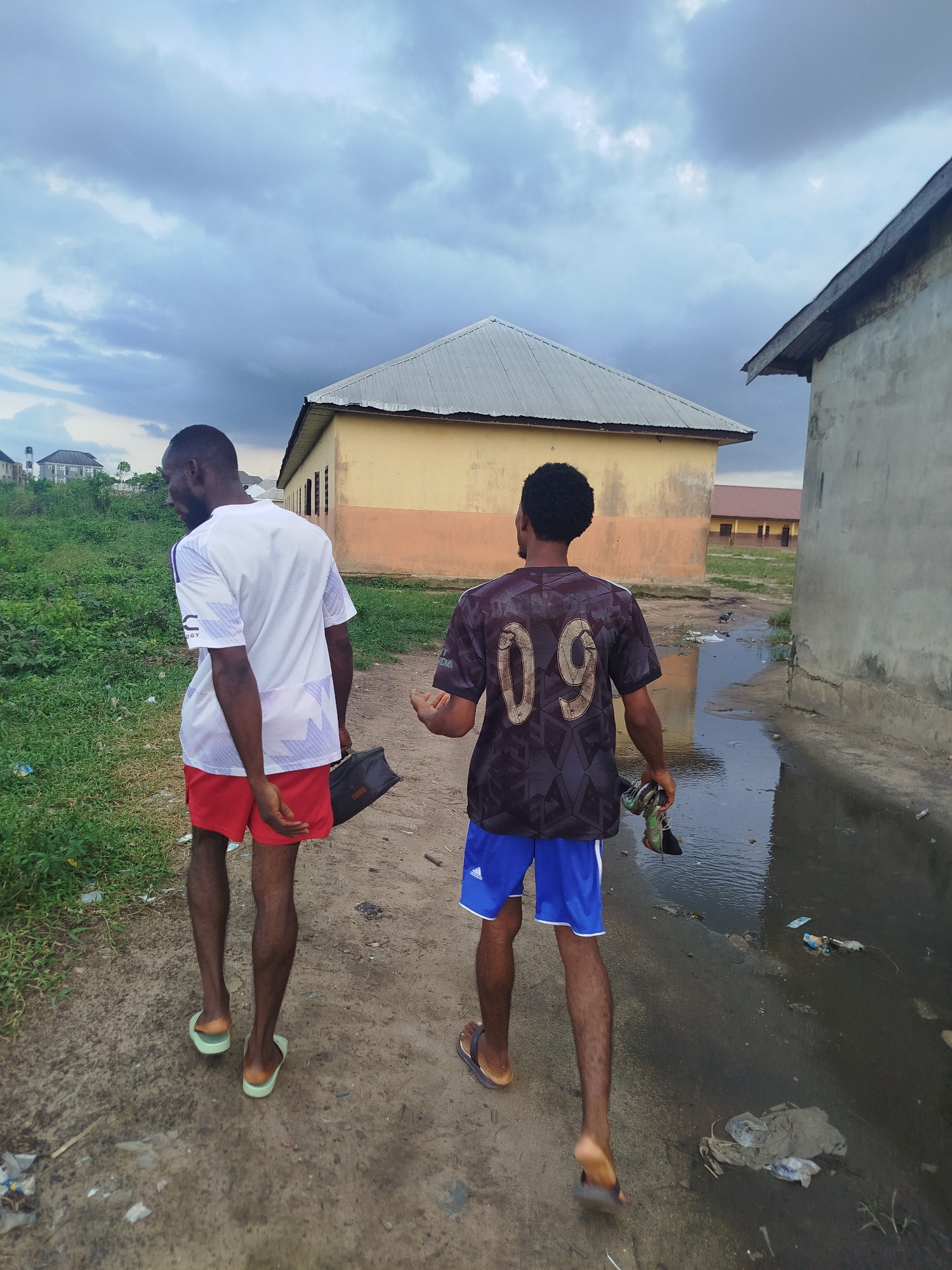Two young men are walking on a dirt path in what appears to be the city of Ikot Mbonde, Nigeria. Both are seen from the back, wearing shorts and flip-flops. The man on the left wears a white t-shirt with red shorts and carries a black bag. The man on the right wears a dark-colored shirt with the number "09" on the back and blue shorts, holding a pair of shoes in his hand. The path is next to a small stream or large puddle filled with trash. Buildings, mostly single-story and weathered, are visible in the background. Overcast sky.