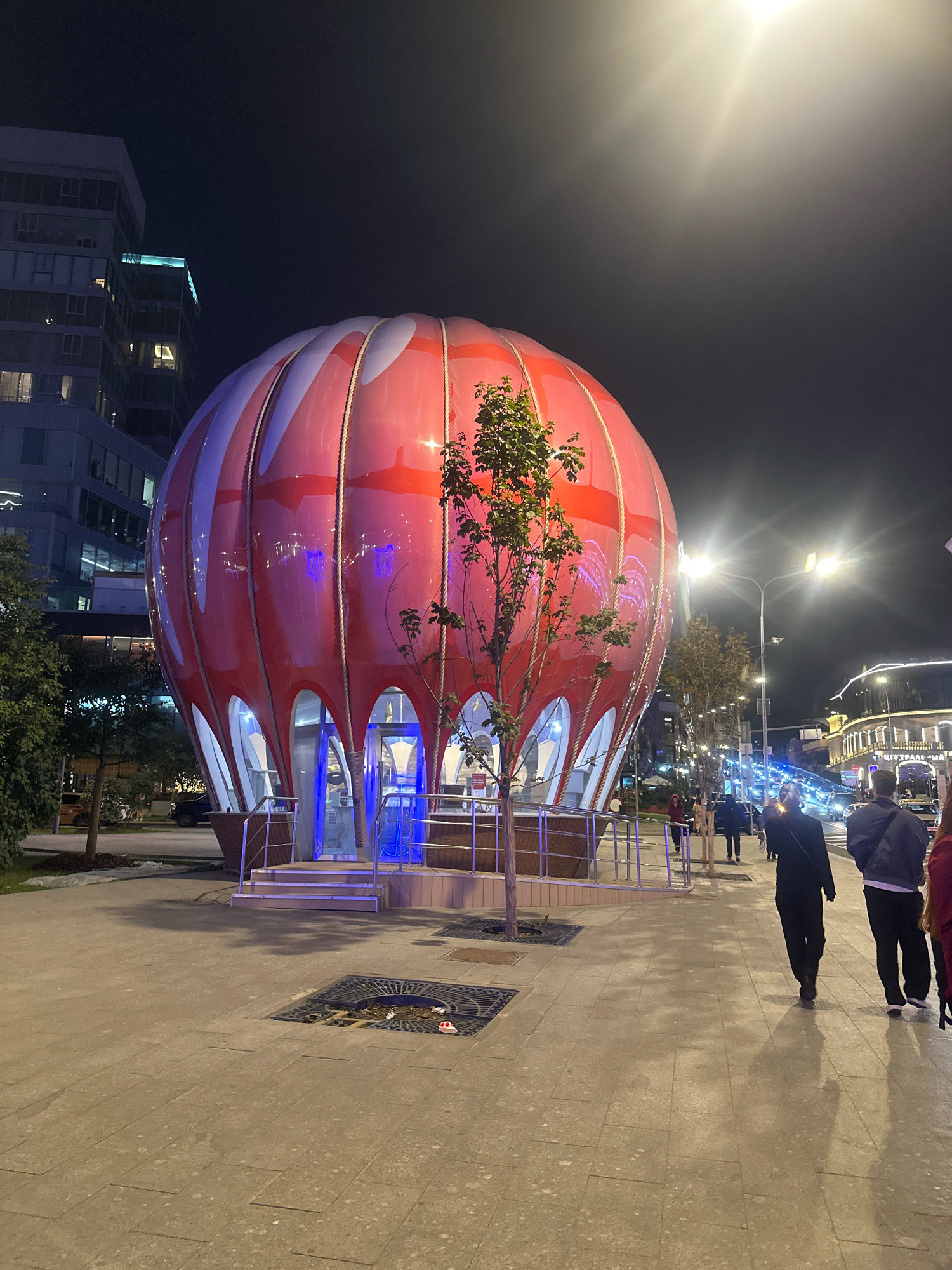 The photograph captures a nighttime scene on a city street in Moscow, Russia, featuring a unique, red and white balloon-shaped structure with arched windows. The structure is illuminated from within with blue light, casting a vibrant glow. In front of it, a young tree stands, its branches reaching towards the balloon.
The street is paved with large, light-colored tiles, and a metal grate covers a drain in the foreground, containing some debris. Several people are walking on the sidewalk. On the left, a modern, multi-story building with numerous windows is visible, partially lit by internal lights and external illumination.
To the right of the balloon structure, the street continues, lined with trees and streetlights. A road with traffic is visible in the background, with car headlights and taillights creating streaks of light. Further down the street, more buildings with illuminated facades are present, suggesting a commercial or entertainment area.
Two individuals are walking away from the camera on the right side of the frame. One person wears a dark jacket and dark pants, while the other is dressed in black. Their shadows are cast long on the paved ground. The overall atmosphere is urban and illuminated, typical of a city at night.