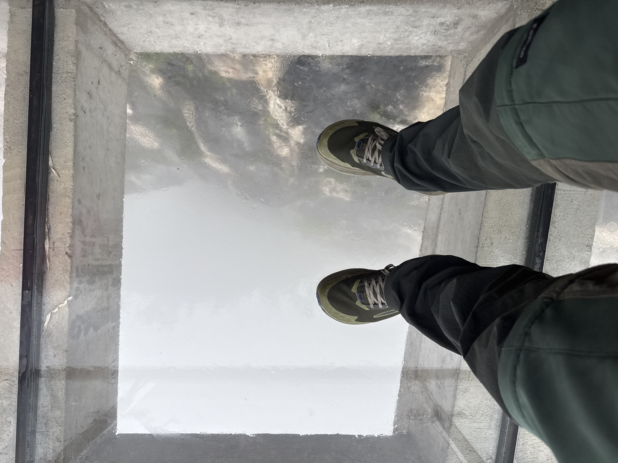 The image shows a first-person perspective from someone standing on a glass-bottomed platform, likely a tourist attraction. The person is wearing gray-green pants and olive green and gray sneakers. Below the glass floor, a cloudy and indistinct view hints at a significant drop, possibly over trees or foliage. The platform is enclosed by gray concrete walls with visible imperfections and textural variations. Based on the prompt, this scene is located in the Yongding District, China.