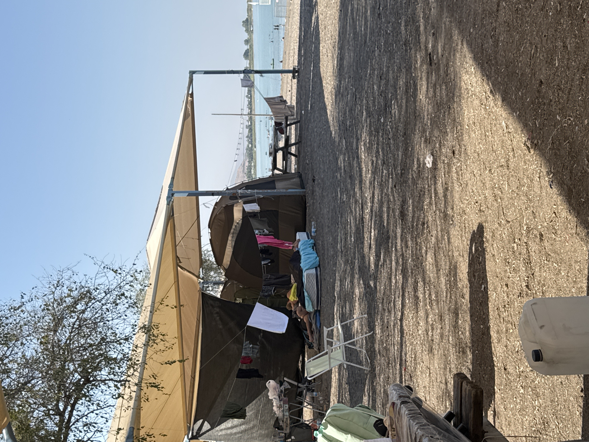 The image depicts a campsite on a beach, likely in the Golan Regional Council, Israel, with a clear blue sky and a calm body of water in the background. A large, tan awning stretches over a camping area, providing shade for tents and a picnic table. Clothes hang on a clothesline strung between supports, some draped over the tents. Two people are lying on mattresses under the awning, seemingly resting or sleeping. A white folding chair is positioned in front of them. To the left, a table with items on it, including a plastic bag, is visible. In the foreground, a wooden picnic table, partially covered with plastic sheeting, is prominent, and a white plastic water container with a black cap sits on the ground nearby. The ground is covered with a mix of sand and wood chips. In the distance, people can be seen swimming and enjoying the water, with hills visible on the horizon.