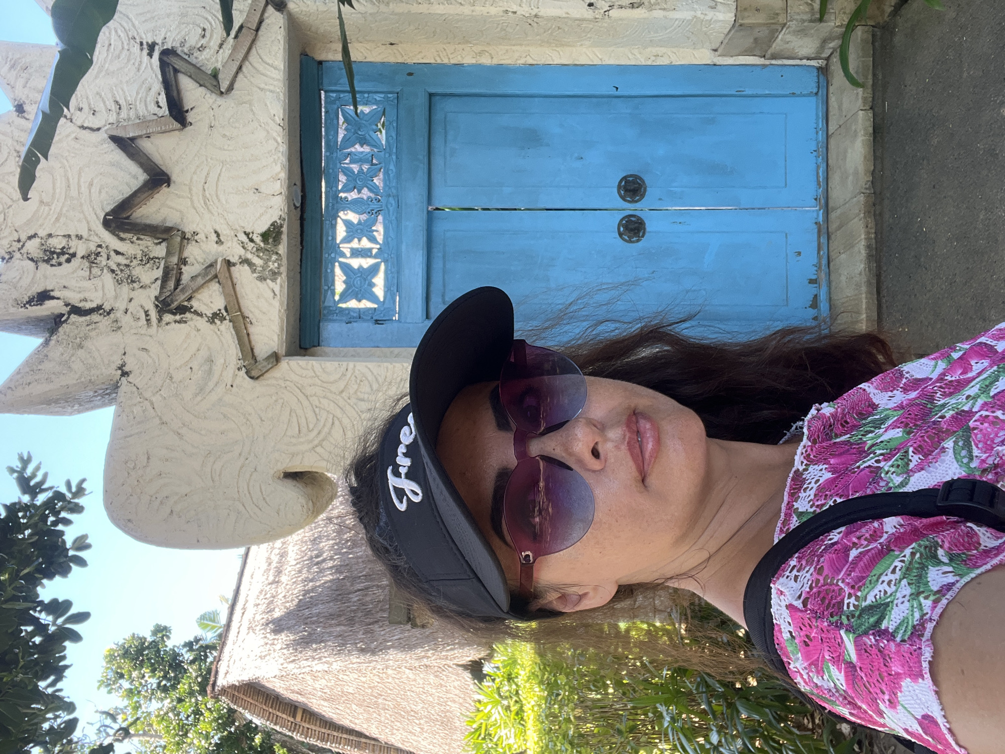 A woman wearing a black visor with white lettering, pink and purple gradient sunglasses, and a pink and white floral top poses for a selfie in front of a blue door. The woman has dark hair and fair skin. The background features a thatched-roof building to the left, lush green foliage, and a white stone structure with decorative carvings above the blue door. The scene appears to be outdoors, likely in a tropical location, such as Kuta Selatan, Indonesia, given the architectural style and vegetation. The blue door is double-paneled with decorative metalwork above and two round, dark metal door handles. The stone structure above the door has a star-like design made of wooden elements. The overall lighting suggests a bright, sunny day.