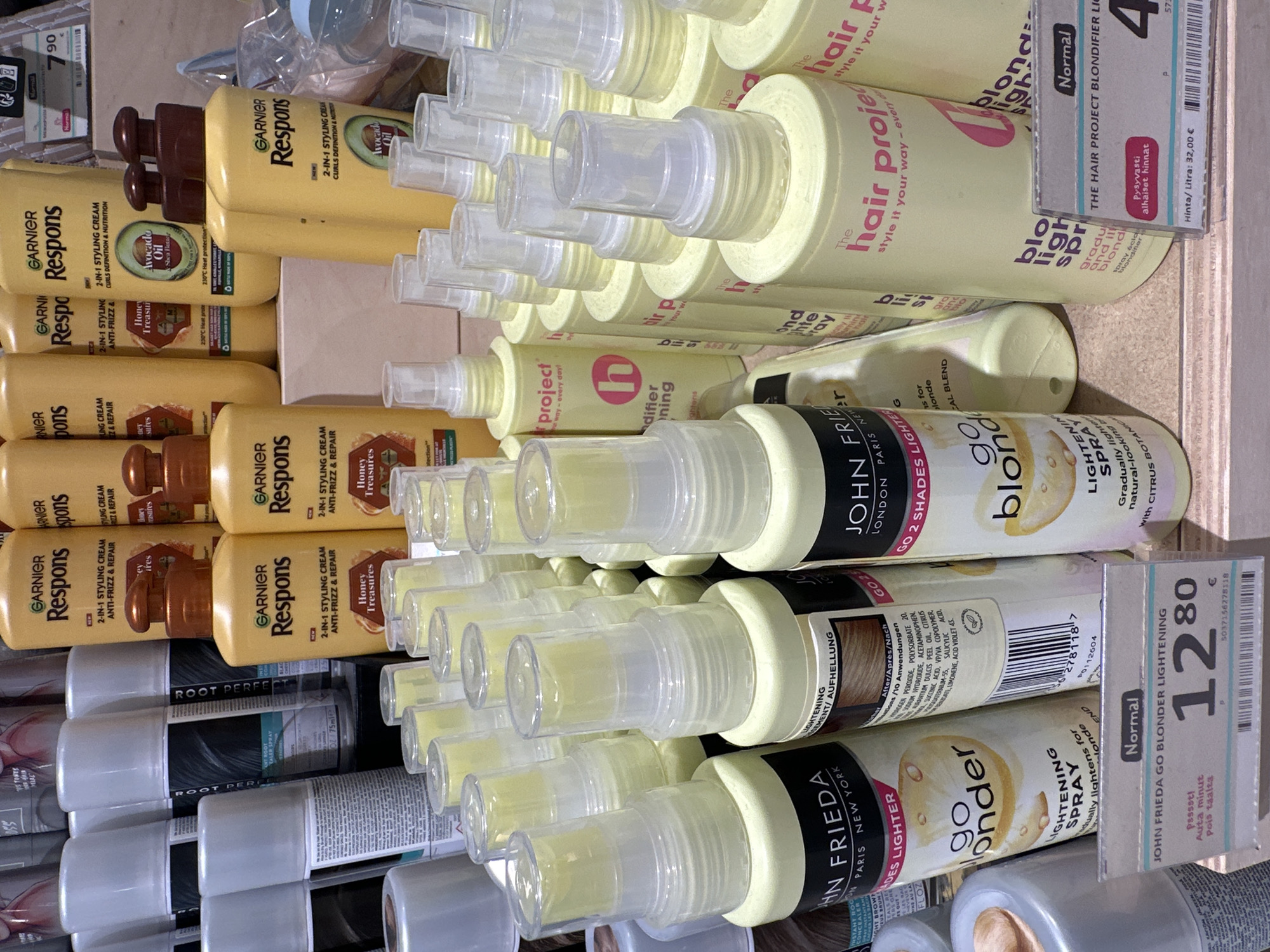 The image displays a retail shelf stocked with various hair care products, primarily in yellow and white packaging. In the foreground, several bottles of "JOHN FRIEDA GO BLONDER LIGHTENING SPRAY" are visible, with one price tag indicating "12.80 €". To the right, "THE HAIR PROJECT BLONDIFIER LIGH" bottles are present, with a price tag showing "4". Further back, numerous bottles of "GARNIER Respons" styling cream are arranged in rows. The overall scene is a well-organized retail display of hair lightening and styling products. The location is an unknown retail environment in Unknown City.