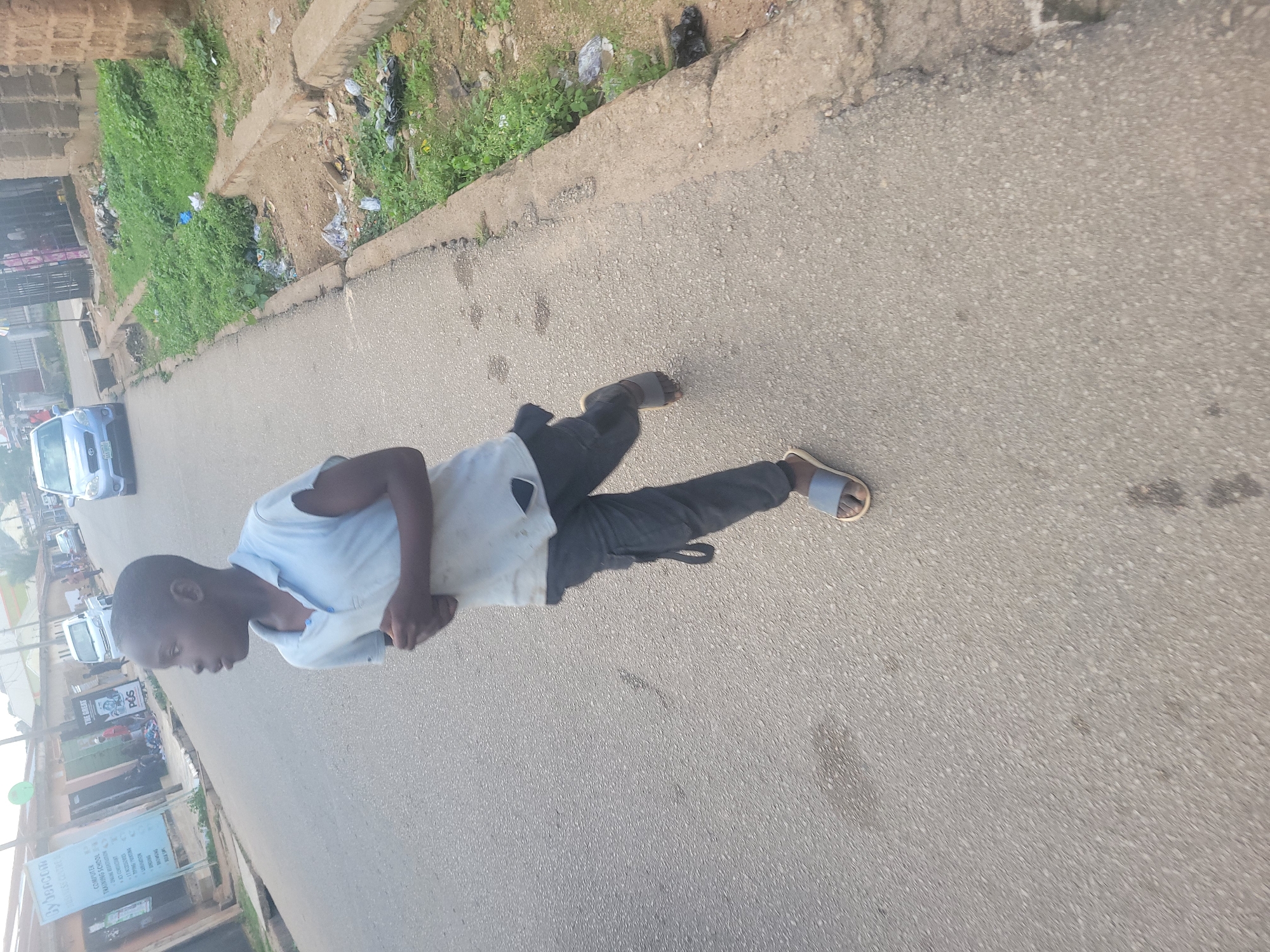 A young boy is running on a paved road in an urban area, possibly Jos, Nigeria. He is wearing a light blue polo shirt and dark pants with pockets. He has on grey sandals. The road is grey asphalt with some patches and stains. To the right of the boy, there is a curb and a ditch filled with green vegetation and some scattered trash. In the background, there are buildings and other vehicles, including a blue car. The lighting suggests it is daytime.