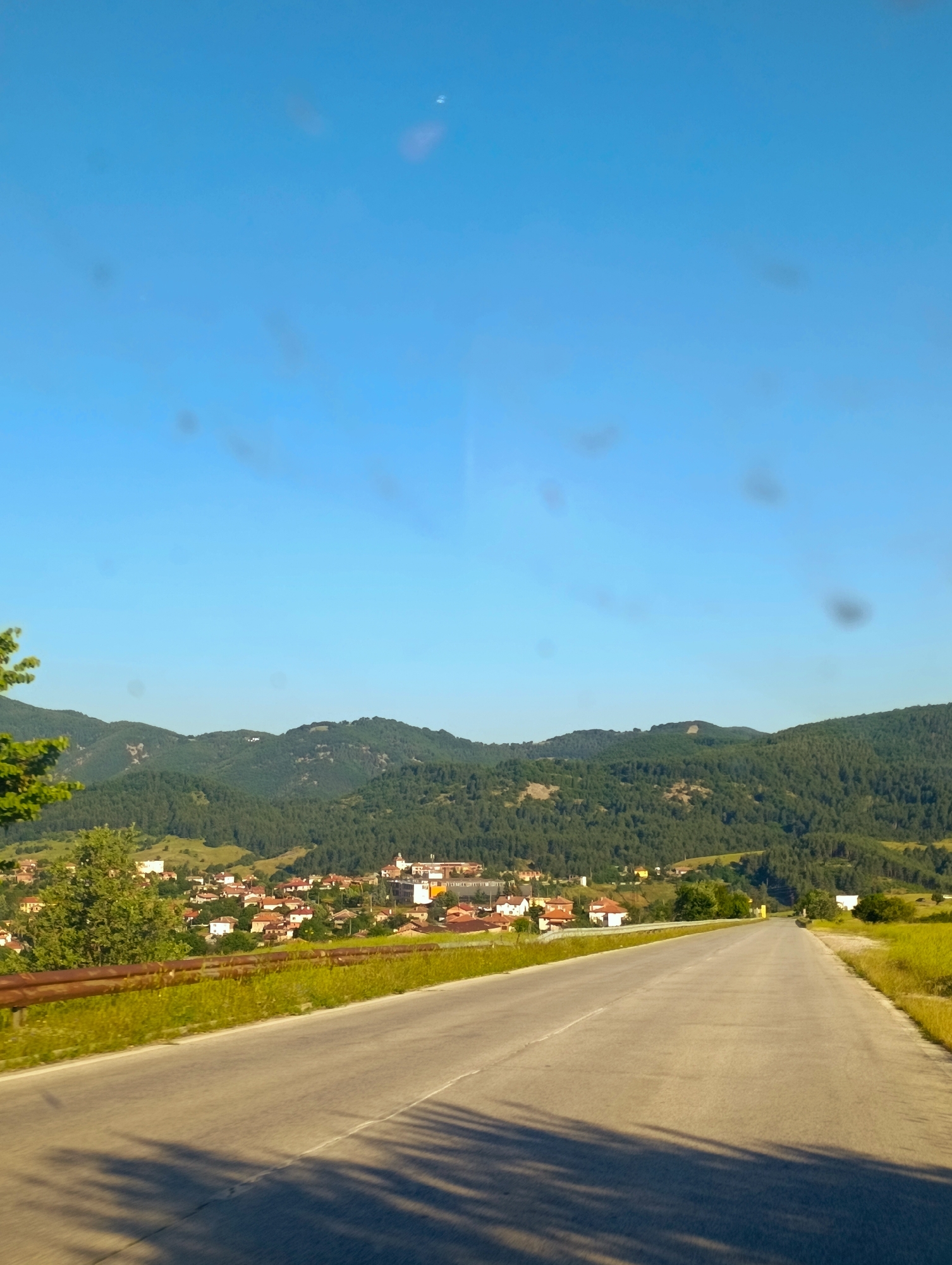 The image captures a rural scene with a road leading towards a village nestled in a valley. The sky is clear and blue, with a few blurry dark spots that appear to be dust or imperfections on the lens. In the foreground, a paved road curves gently to the right, casting long shadows from an unseen object. A rusty metal guardrail runs alongside the road, bordered by green grass.
Beyond the road, a village with red-tiled roofs and white and yellow buildings is visible. The village is situated at the foot of a densely forested mountain range. The mountains are covered in dark green trees, with some lighter patches indicating clearings or different vegetation. The overall impression is one of a peaceful, sunlit rural landscape. The specific location in the city of Komuniga, Bulgaria, is indicated by the provided context, suggesting the scene depicts a part of this Bulgarian settlement and its surrounding natural environment. There are no people or animals visible in the frame.