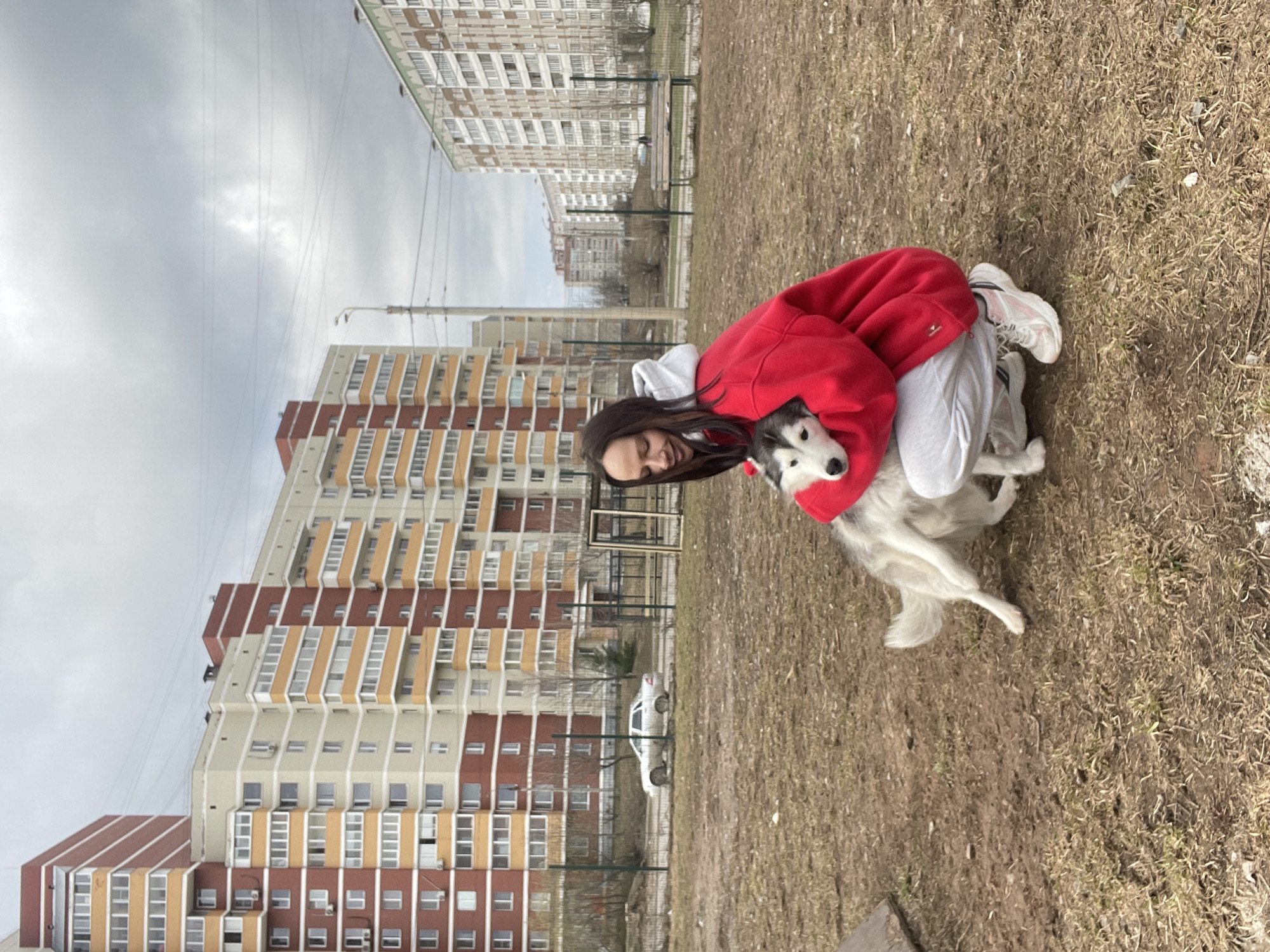 A young woman is captured in a heartwarming moment, embracing a black and white husky dog in an urban outdoor setting in Izhevsk, Russia. The woman, with long dark hair, is smiling and wears a red hooded sweatshirt, light grey sweatpants, and white sneakers. She is kneeling or crouching on dry, sparse grass and dirt, holding the husky close to her, with the dog's head resting on her shoulder. The dog appears calm and content.
The background features several large, multi-story apartment buildings with distinctive white, brown/red, and orange sections, characteristic of residential architecture in the region. Overhead utility wires are visible against a uniformly overcast, grey sky. A dark metal fence separates the immediate grassy area from a paved surface behind it, where a white car is parked. Another wooden bench is partially visible behind the fence. The dry ground suggests the season is either late winter or early spring. The scene conveys a sense of affection and companionship on a cloudy daytime. No visible text is present in the image.