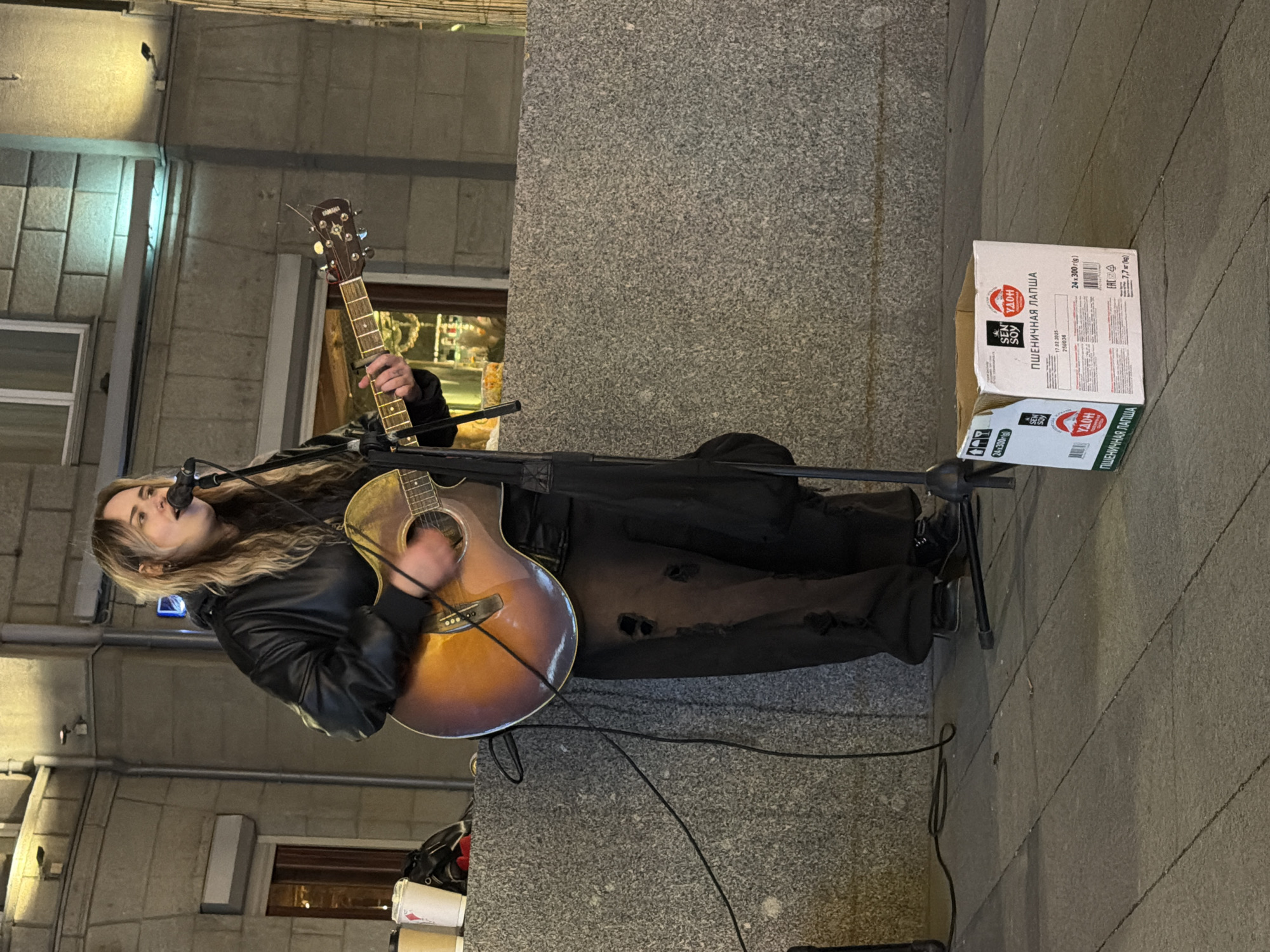 The image shows a woman performing as a street musician in what is likely the city of Moscow, Russia, based on the Cyrillic writing on a box next to her. She is playing an acoustic guitar and singing into a microphone. She wears a black leather jacket and dark pants with some rips or distressed elements. A black bag is near her feet. Her blonde hair is long and loose.
A cardboard box with Cyrillic text sits on the pavement beside her. The text includes "SEN SOY" and "ПШЕНИЧНАЯ ЛАПША" which translates to "WHEAT NOODLES". A microphone stand holds the microphone in front of her. The scene appears to be outdoors in an urban area. Buildings with windows are visible in the background. The lighting suggests it is evening or night. The pavement appears to be made of large, gray stone slabs.