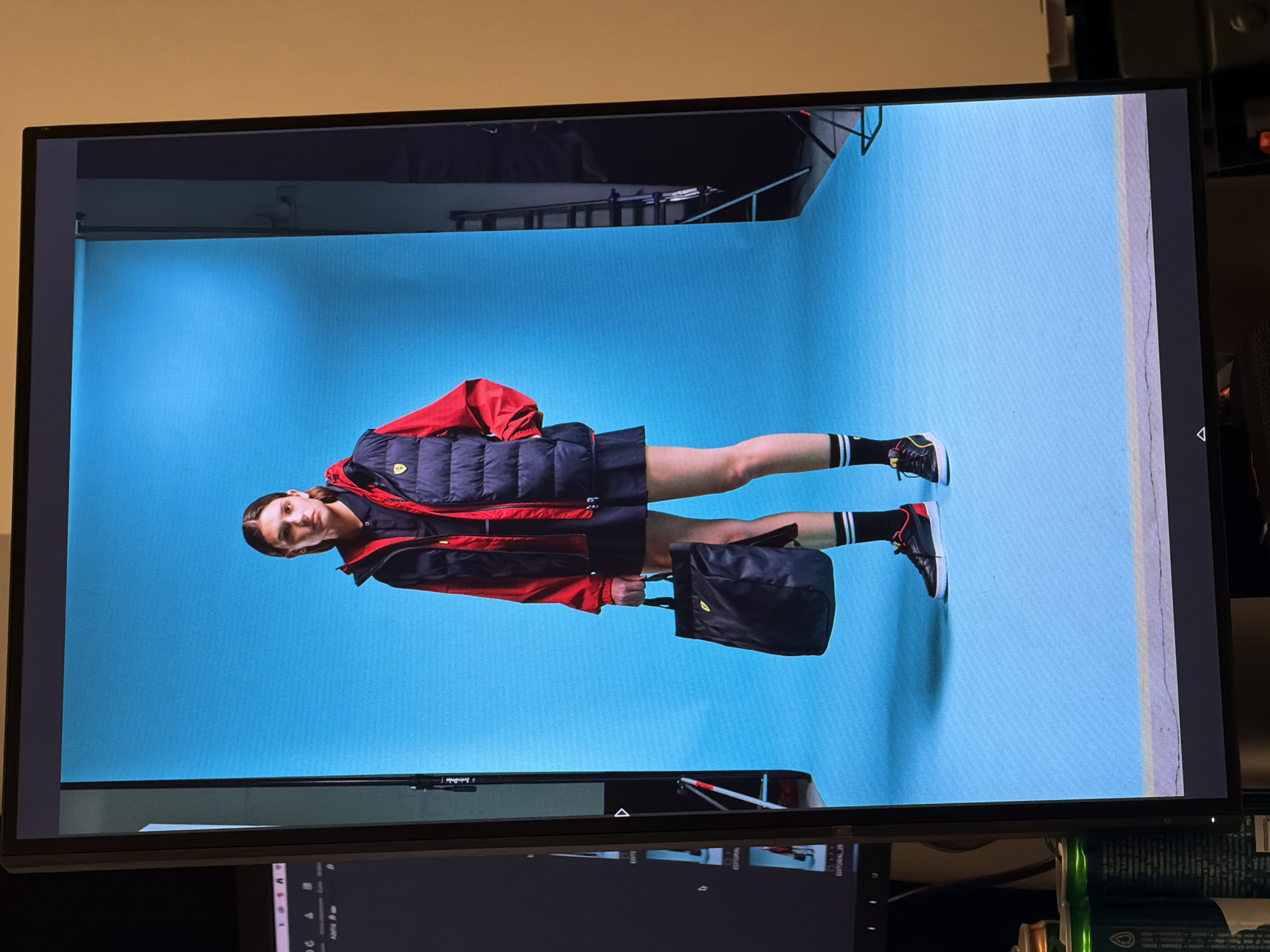 The image shows a monitor displaying a fashion photograph. The photograph features a model standing on a light blue backdrop, possibly in a studio setting. The model is wearing a red and navy puffer jacket, a navy skirt, black and white striped socks, and red and black sneakers. She is holding a black tote bag. The background suggests an indoor studio environment with lighting equipment visible above. It cannot be ascertained that this image was taken in Milan, Italy.