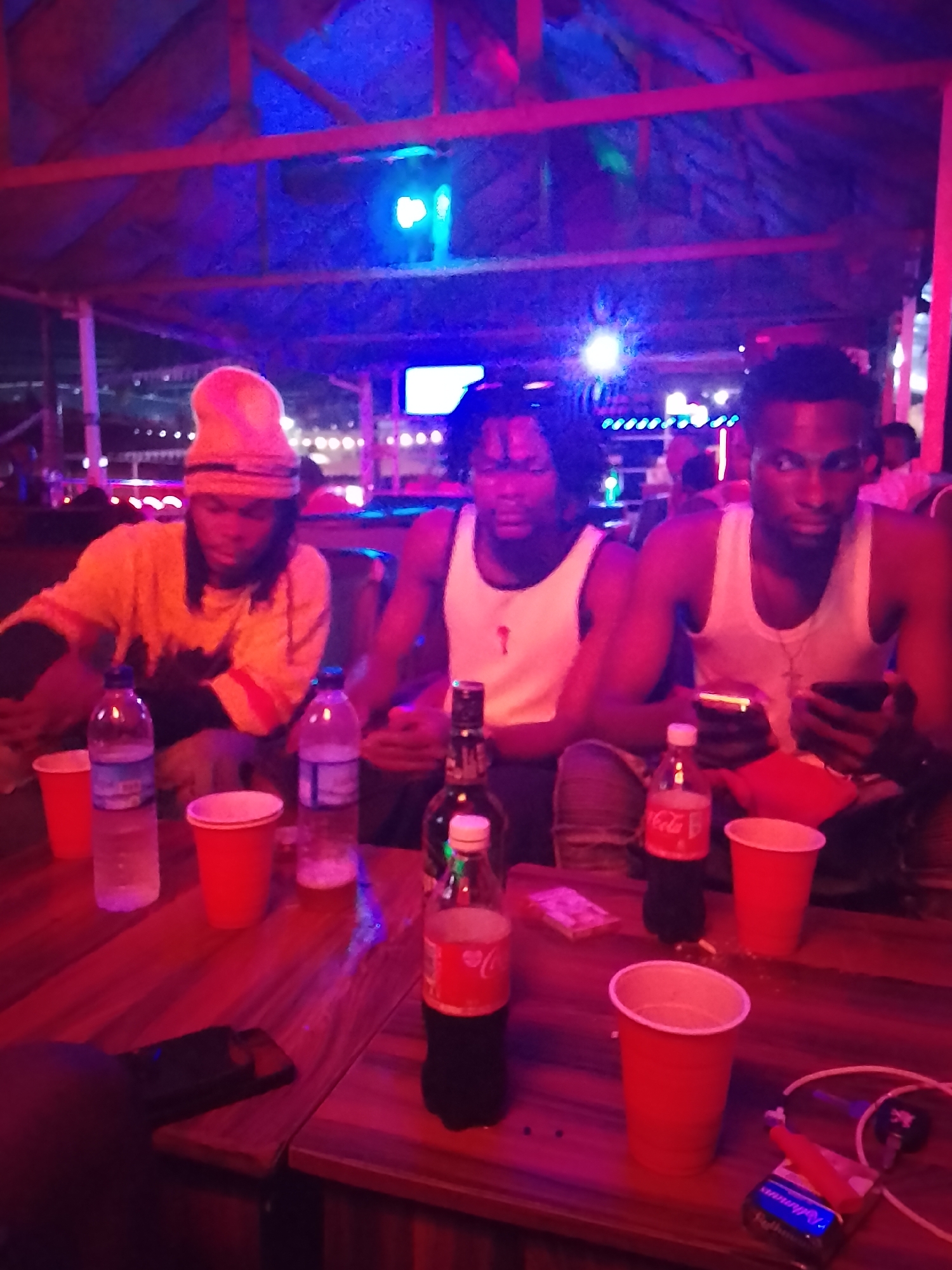 The image shows three men sitting at a wooden table indoors. The man on the left wears a yellow shirt and hat. The other two men wear white tank tops. On the table are bottles of water, Coca-Cola, a liquor bottle, red plastic cups, cigarettes, and phones. The location is a bar, identified by the festive lighting. The city is Unknown, Unknown.