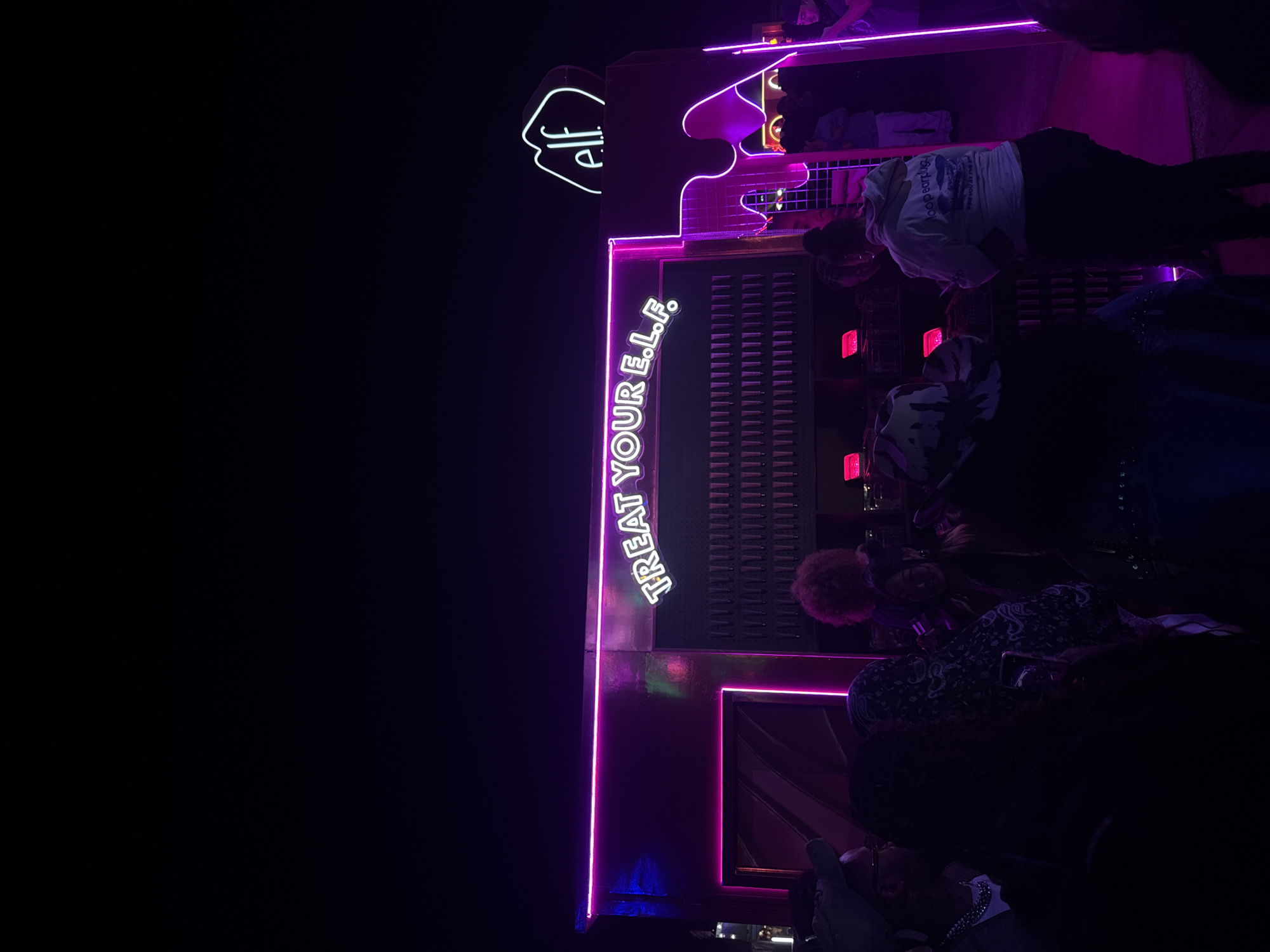 The image depicts a nighttime scene in Thermal, United States of America, featuring a display or structure with neon lighting. The text "TREAT YOUR E.L.F." is prominently displayed vertically with neon letters. The logo of "e.l.f." is also visible in neon. Several people are present, though their faces are mostly obscured by darkness and the orientation of the photo. Some individuals appear to be interacting with the display. The environment is dark, with the neon lights providing the primary illumination. The background is indistinct, suggesting an outdoor location.