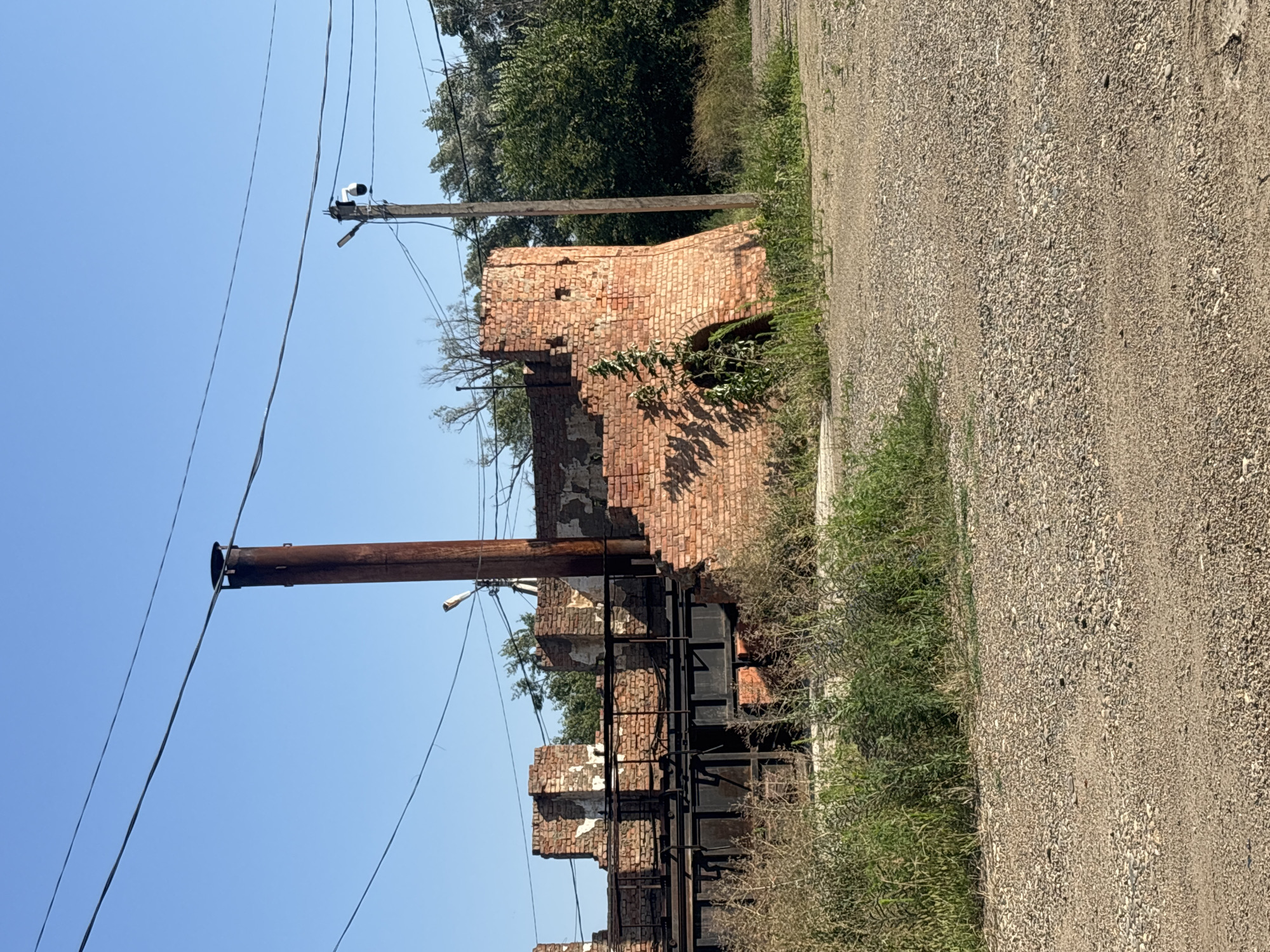 An outdoor, eye-level, medium shot captures the ruins of a brick building under a clear blue sky. The building, located in Arkhipovskoe, Russia, appears to be an old industrial structure, possibly a brickworks, with a large, rusty metal chimney rising from its center. The brick walls are damaged and weathered, with patches of white plaster visible. To the right of the chimney, a section of the building has a rounded archway. A tall, wooden utility pole stands near the ruined structure, with a security camera and a streetlight attached to it. Overhead, power lines crisscross the sky. The foreground is dominated by a gravelly dirt road, with overgrown green vegetation on either side. The overall scene conveys a sense of abandonment and decay.