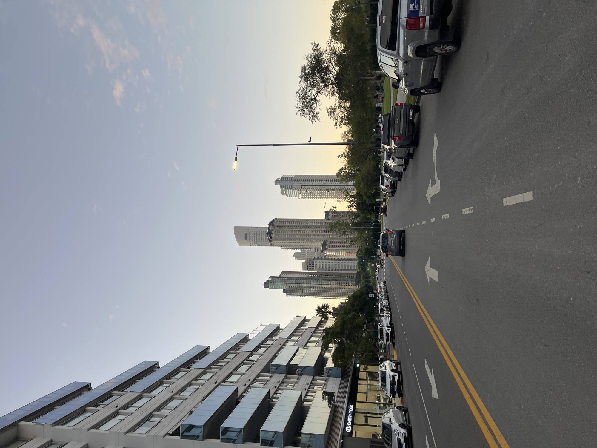 The image captures a wide street in Buenos Aires, Argentina, lined with parked vehicles on both sides. The perspective aligns along the street, emphasizing its length and leading the eye towards a distant cluster of skyscrapers. A gray car is traveling down the street. Buildings with distinct modern architecture, featuring multiple stories and visible windows or balconies, dominate the foreground. Street lights are interspersed along the road. The sky above is light, suggesting either early morning or late afternoon. The scene portrays an urban environment with a mix of residential and commercial buildings.