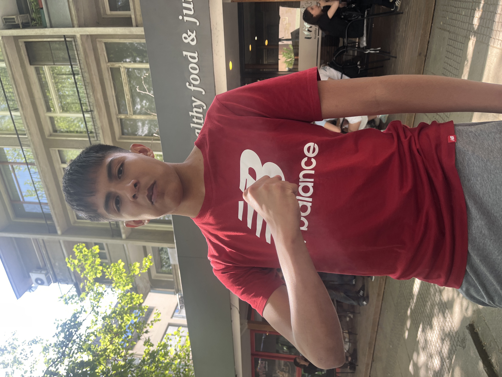 A young male stands in front of a building with a sign that reads "Healthy food & ju" in Plovdiv, Bulgaria. The building appears to be a restaurant or cafe. The man is wearing a red New Balance t-shirt and gray pants. He is making a fist with one hand. Behind him, other people are sitting at tables. The weather appears to be sunny, with green trees visible in the background. The architecture suggests a modern building.