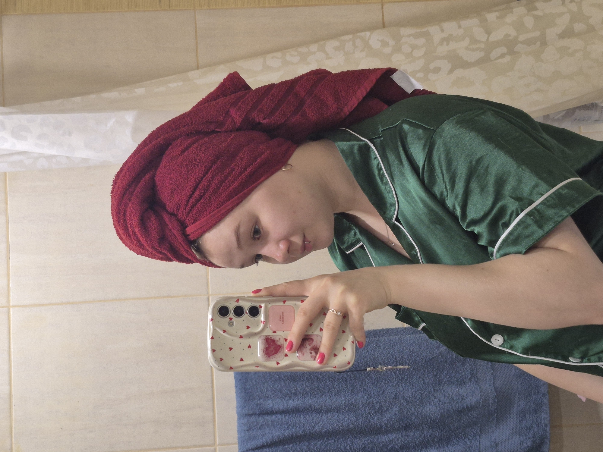 The image displays a young person taking a selfie in what appears to be a bathroom mirror. The subject, with fair skin, is wearing dark green satin pajamas with white piping and has their head wrapped in a deep red towel, suggesting they have recently washed their hair or showered.
They hold a white smartphone with a decorative case featuring numerous small red hearts and several larger pink, heart-shaped or square embellishments. On one of the pink rectangular sections of the phone case, reversed text is visible, which appears to read "Adelante" above a partially obscured word or number. The person is wearing a silver-colored ring on their right ring finger and a thin silver necklace. A small, metallic piercing is also visible near their lower lip.
The background shows light-colored, possibly beige, tiled walls, typical of a bathroom. A white shower curtain with a subtle, light-colored pattern (resembling leopard print) is visible in the upper left corner. A blue towel is draped below the person's arm and phone. The subject's expression is neutral and focused on the phone screen. The overall scene suggests an intimate moment of self-care or routine. The attire (pajamas) implies either morning or evening. The provided location context is Chaikovsky, Russia.