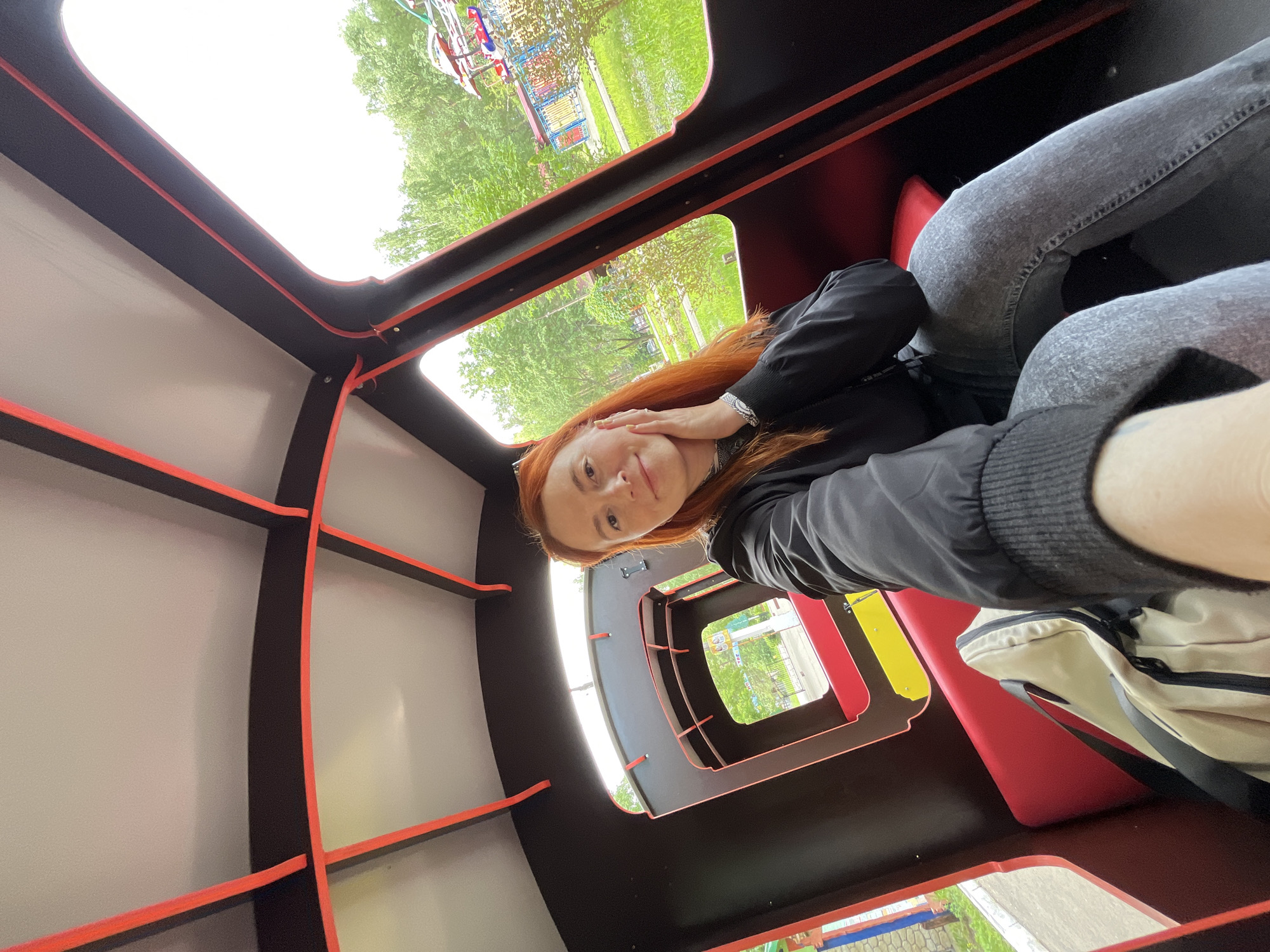 A ginger-haired woman takes a selfie inside a red and black train car ride within an amusement park in Chaikovsky, Russia. She is positioned slightly to the right of the frame, smiling faintly, with her left hand touching her cheek. She wears a black jacket with ribbed cuffs, a patterned top, and light grey jeans. A beige backpack rests on the red seat beside her. The train car's interior features black walls, a light grey ceiling, and bright red trim along the structural elements and window frames. Through the windows, a lush green park environment is visible, with numerous trees, grass, and several colorful amusement park rides, including one with umbrella-like structures and another with vibrant blue, red, and yellow panels. A paved path runs alongside the train car on the left.