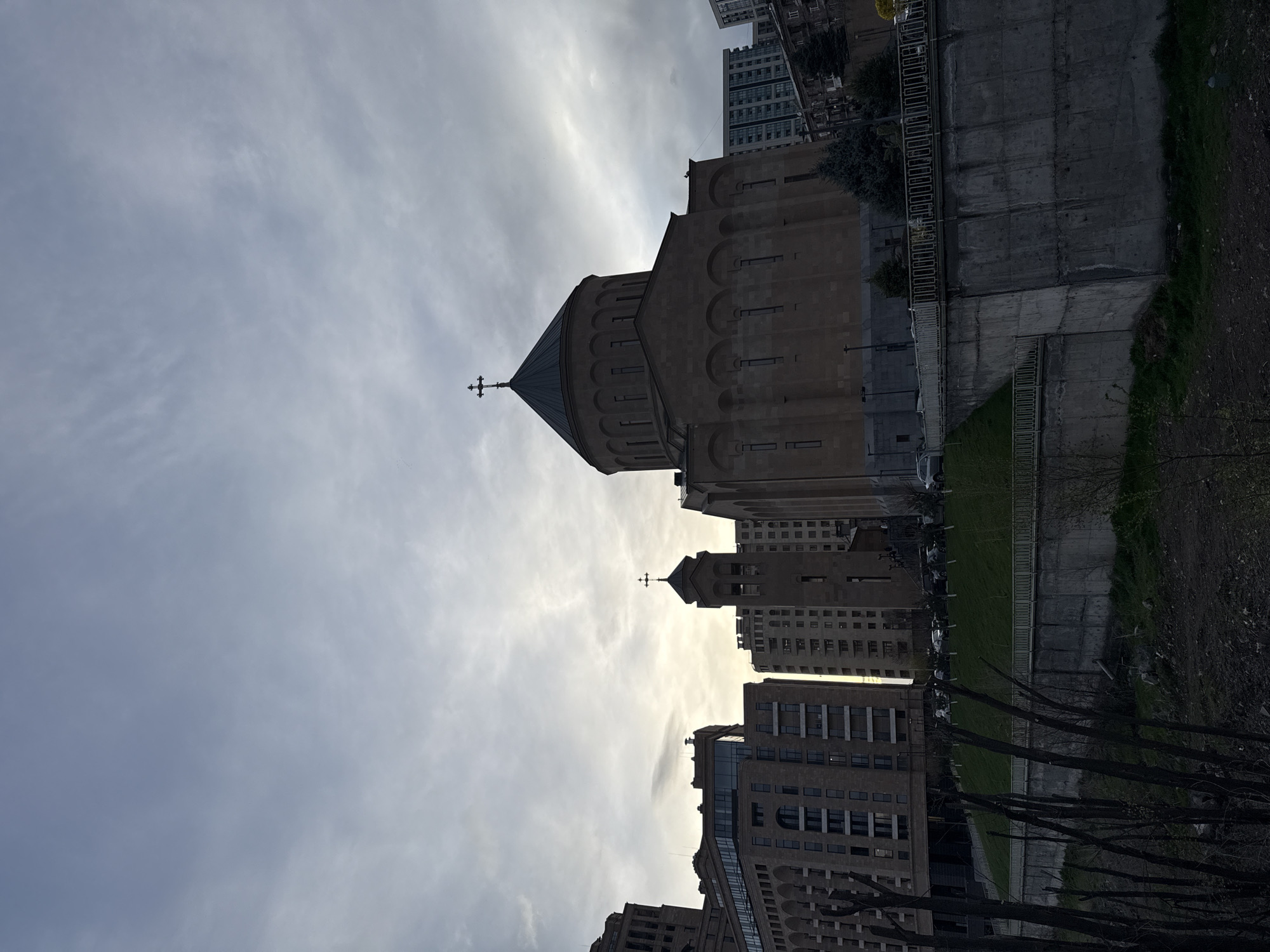 The image depicts an urban landscape in Yerevan, Armenia. The primary subject is the Saint Anna Church, a prominent religious building with a cone-shaped roof topped with a cross. The church is surrounded by modern high-rise buildings. A concrete wall and a strip of green vegetation are visible along one side of the church. The sky is overcast. The angle of the shot gives a low perspective, looking upwards at the church and the surrounding buildings. A secondary church is between the apartment blocks. Some trees with bare branches are in the foreground.