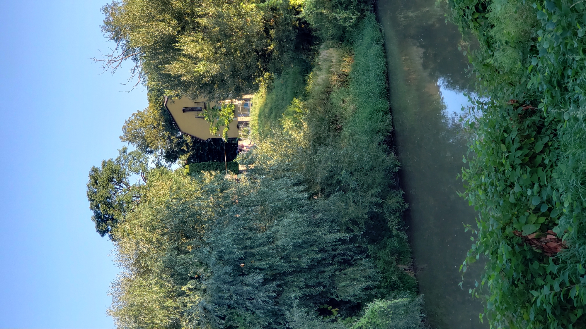 The image depicts a serene natural scene with a yellow house situated on a grassy hill, partially obscured by lush green foliage. The house, with a dark-tiled roof and a single window visible on the upper floor, has a small balcony. A fence surrounds a portion of the property. In the foreground, a body of calm, dark water reflects the sky and surrounding greenery. Dense vegetation lines the bank of the water. Tall trees with dense canopies dominate the background, with a clear blue sky above. Two indistinct figures can be observed on the hill near the house. The overall impression is one of a tranquil summer day in a semi-urban or rural setting.