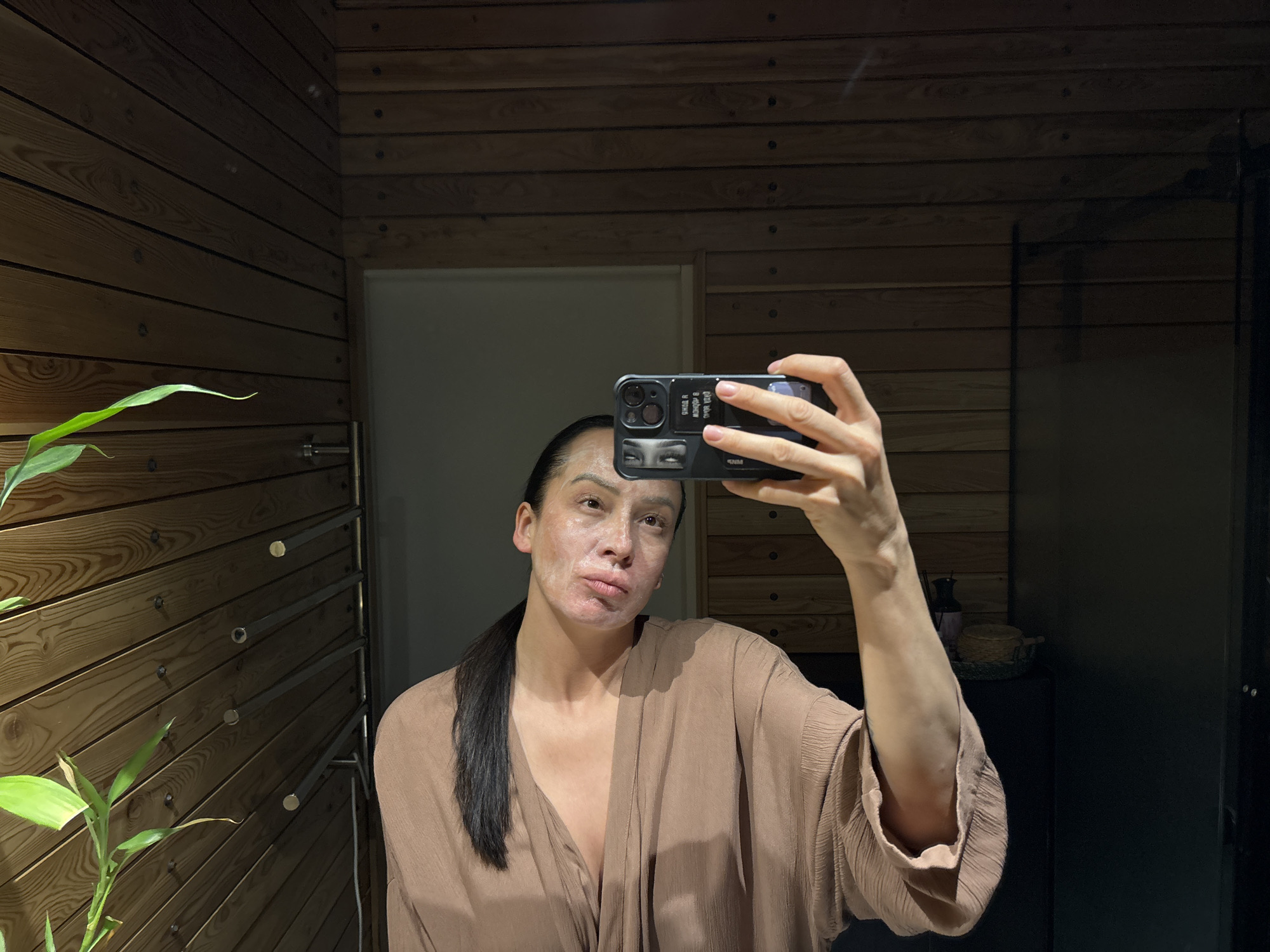 A person is taking a selfie in a bathroom. The bathroom has wooden paneling on the walls and a white door. The person has a face mask on and is wearing a brown robe. They are holding a smartphone with a black case. A plant is in the lower left corner of the image. The location may be in Chaikovsky, Russia.
