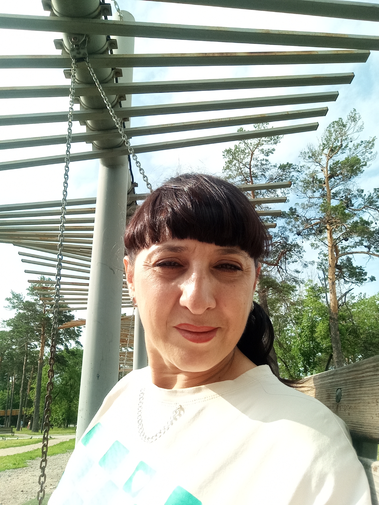 The image shows a medium shot of a woman with dark hair and bangs, wearing a light-colored t-shirt with a geometric pattern and a silver necklace. She is outdoors, with a sky and trees in the background. To her left, there is a structure with metal bars and chains, possibly part of a canopy or shelter. The woman is looking towards the camera with a slight smile. The lighting suggests it is daytime, with sunlight casting shadows. The overall setting appears to be a park or outdoor recreational area. It is not possible to determine the location in the city of Belogorsk, Russia from the image alone.