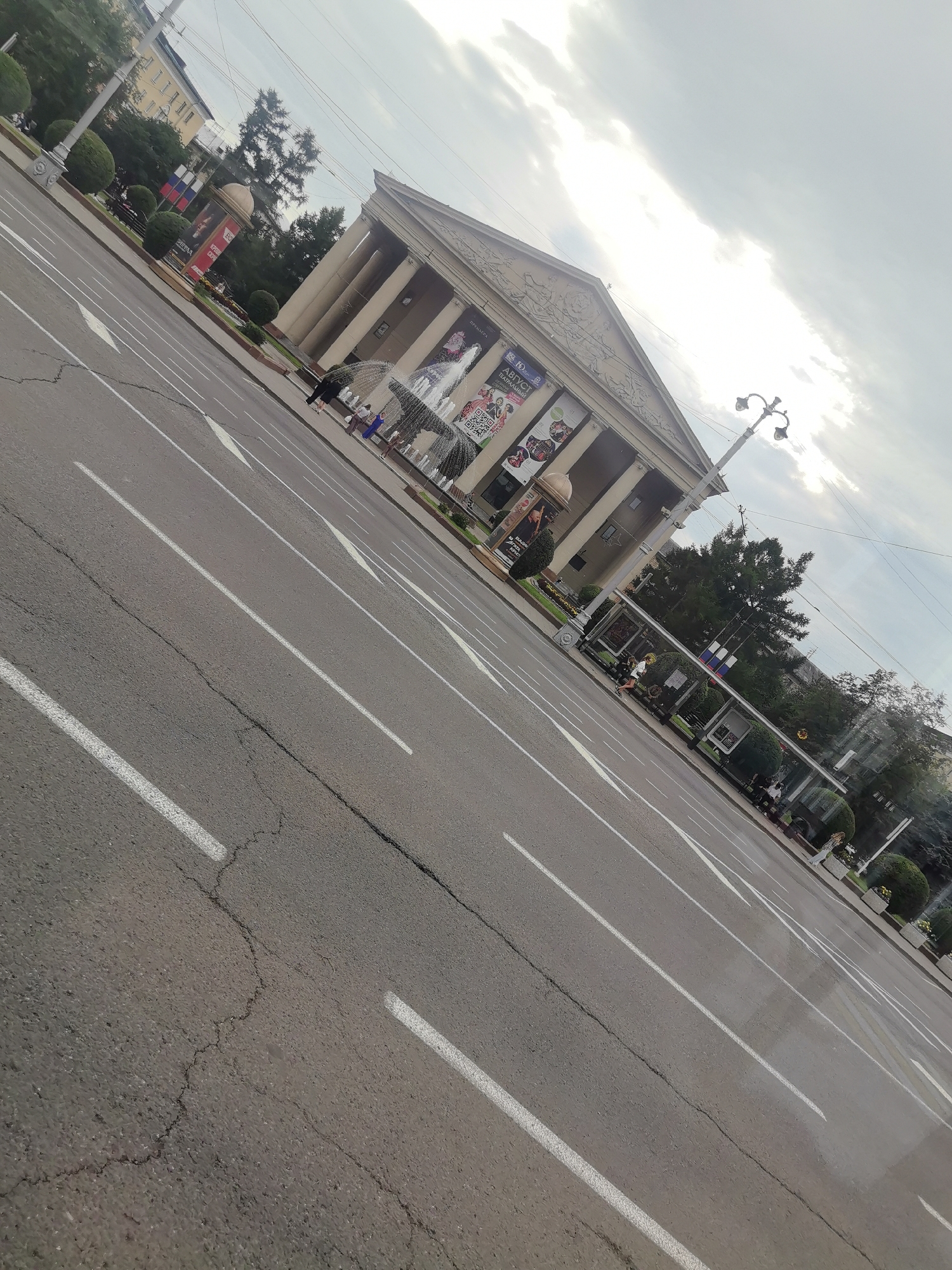 The image shows a wide, cracked asphalt road with white lane markings. The road is angled diagonally across the frame, suggesting a tilted perspective or a moving vehicle. On the right side of the road, there is a sidewalk and a bus stop shelter with people waiting. Beyond the sidewalk, there are trees and a large building with classical architecture, featuring columns and pediments. Several banners are hung on the building, some displaying text and images. A fountain is visible in front of the building. The sky above is cloudy, with patches of bright light suggesting the sun breaking through. The scene appears to be a public space in a city, likely a square or main street.
