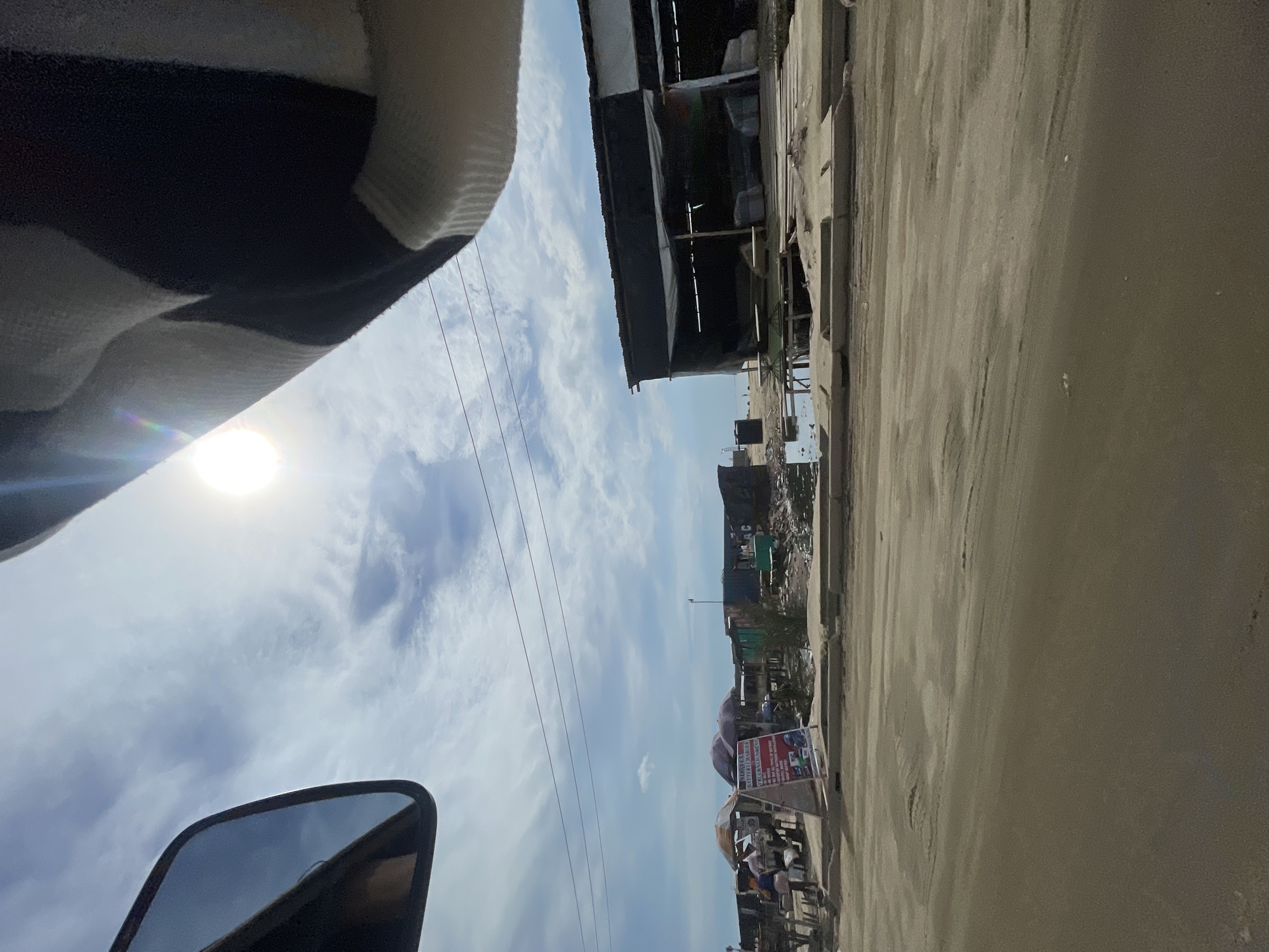 A photograph taken from within a vehicle shows a roadside scene in Ado, Nigeria. The sun is high in the sky, partially obscured by clouds, with overhead power lines visible. The road is dusty and textured. Buildings, including a shed-like structure with a dark roof, line the side of the road. A sign with red and white text, possibly advertising food or produce, stands near a cluster of people. The side mirror of the vehicle is visible.
