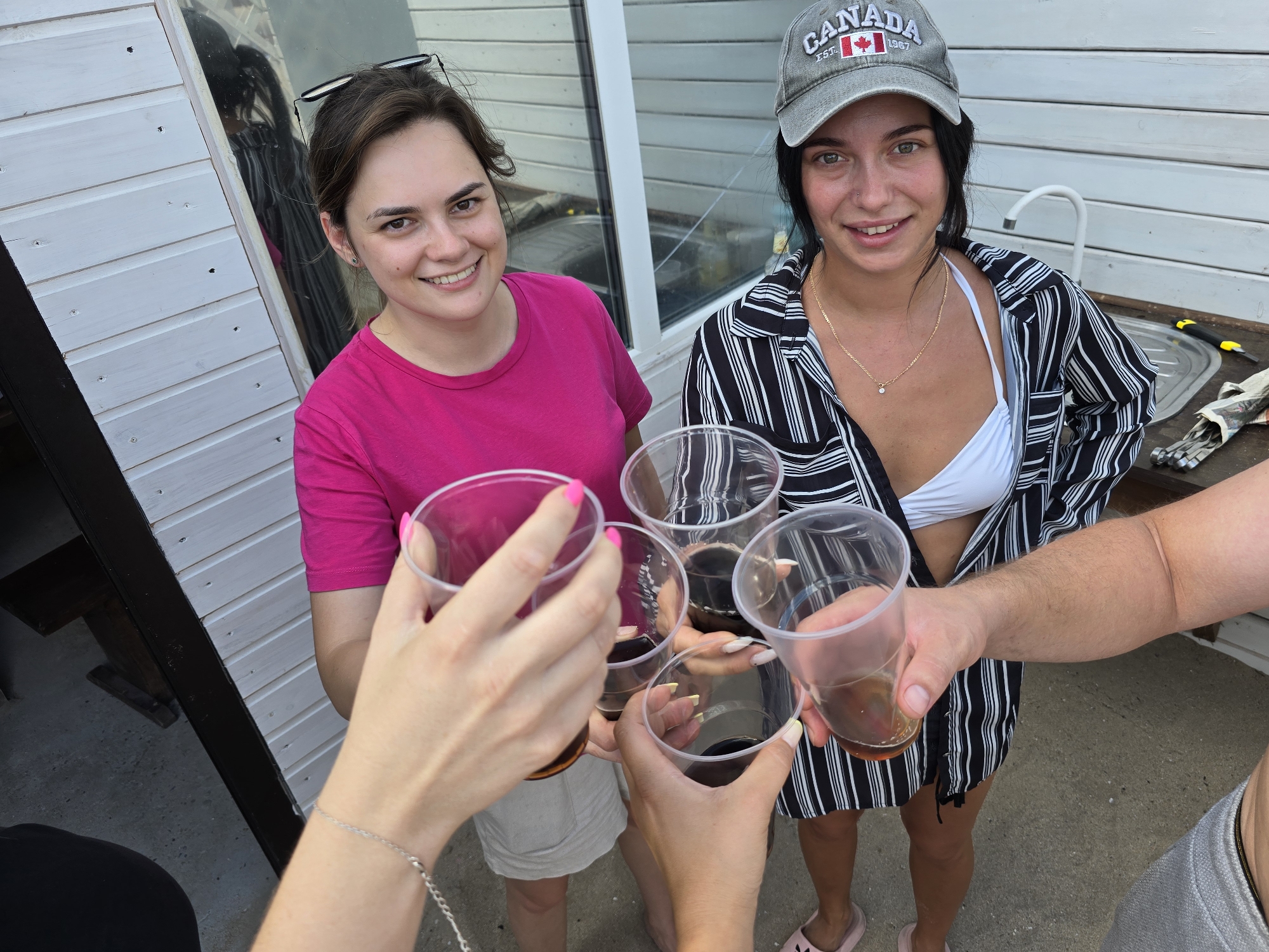 In Yevpatoriya, Ukraine, a group of four people is captured in a candid outdoor shot, appearing to be mid-toast. The setting is rustic, with a white wooden structure behind them and a concrete ground.
On the left, a woman with dark brown hair tied back and glasses perched on her head smiles brightly, wearing a vibrant pink t-shirt. To her right, another woman, wearing a grey "Canada" baseball cap, a black and white striped open shirt over a white bikini top, and a delicate gold necklace, also smiles.
The focus is on their hands, raised in a toast with clear plastic cups filled with amber-colored liquid. The hands are adorned with nail polish and jewelry, suggesting a casual, social gathering. One hand on the far left has a silver bracelet.
The environment appears to be an outdoor area, possibly a backyard or patio, with a sink and a tool visible on a counter to the right, hinting at a functional space that may also serve for leisure. The lighting suggests it is daytime, with a slightly overcast sky.